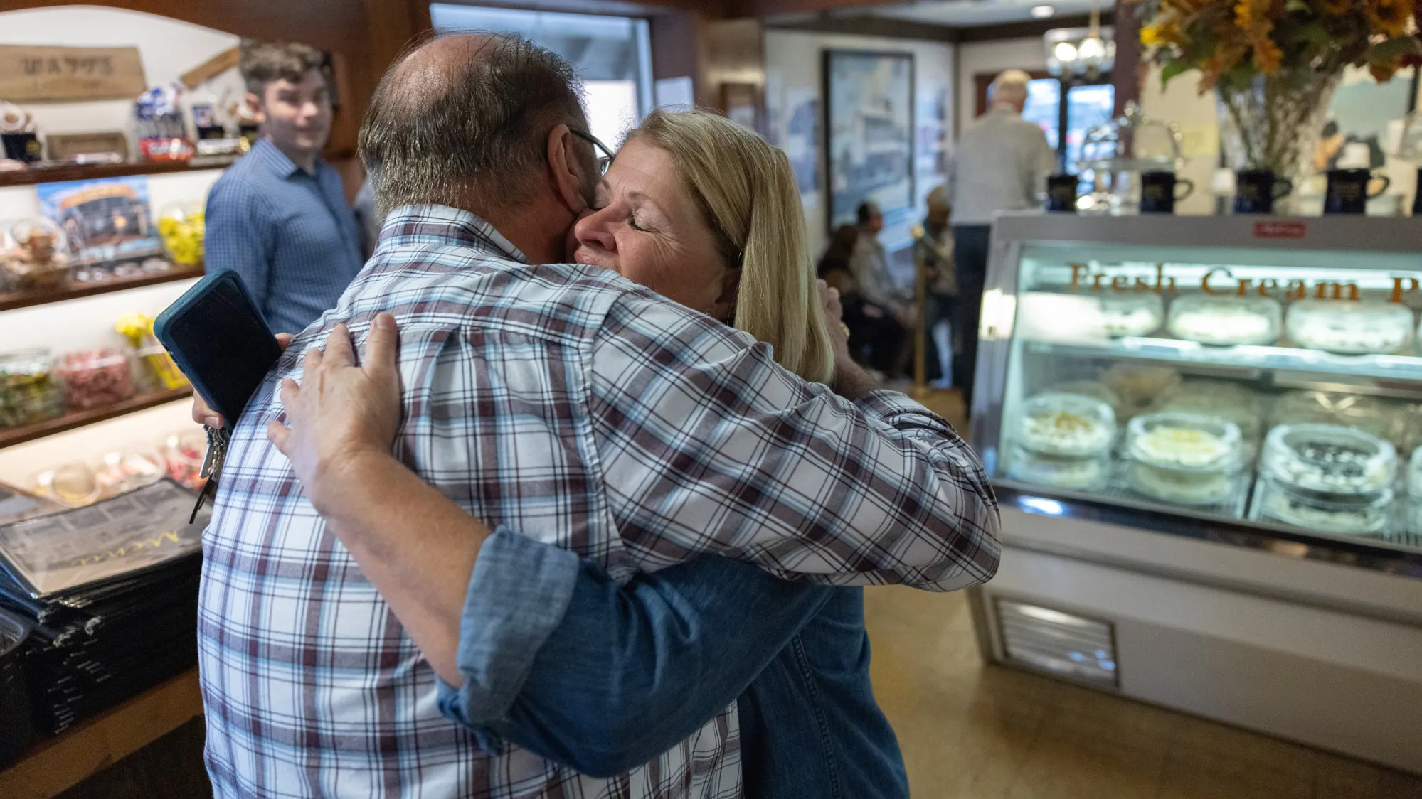 In a restaurant room (possibly an entry) lined with cases of pies and other food items for sale, a woman in a denim shirt hugs a man wearing a plaid shirt. 
