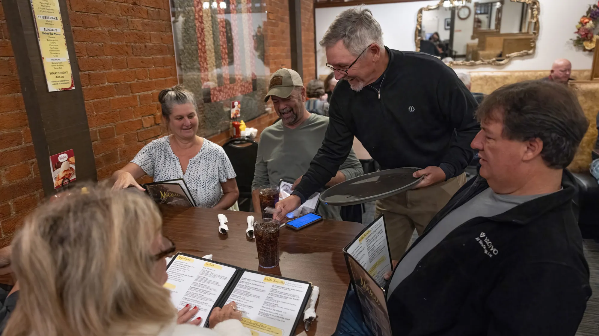 Diners with menus laugh as a man holding a tray sets down their sodas. An exposed-brick wall is next to them and the table is cozily-sized. 