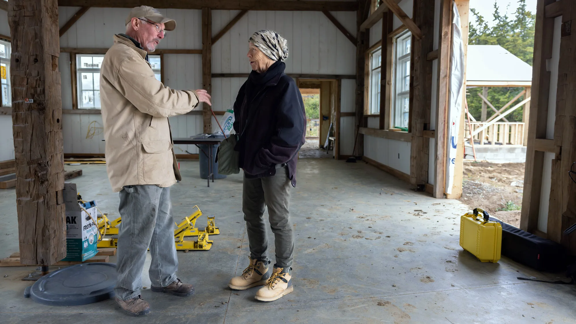 Inside the barn that is under construction, Doug Morgan gestures as he talks with an older woman wearing a hat and fall windbreaker. Around them, there is some workers’ equipment, the floor is marked by muddy boot prints and the walls are primered a bright white. 