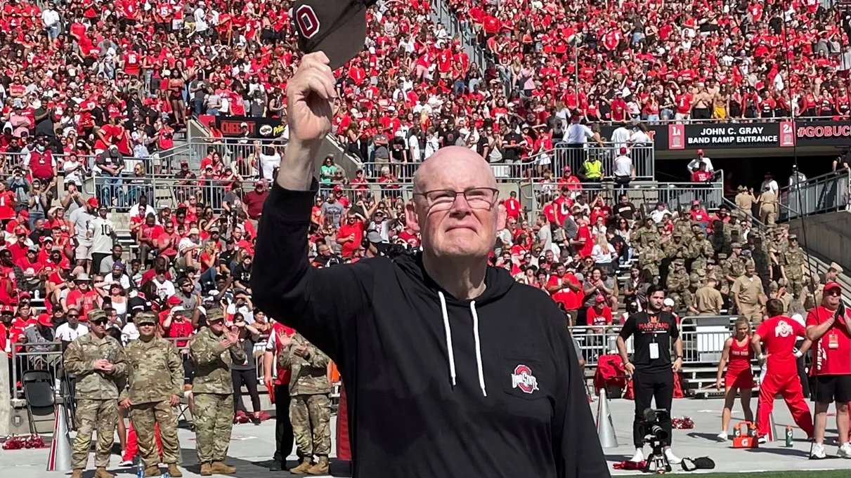 Standing on the field inside Ohio Stadium, Daniel Connor, an older white man dressed in matching pants and sweatshirt—with an Ohio State logo—waves his baseball cap in the air as he greets the crowd at a football game.