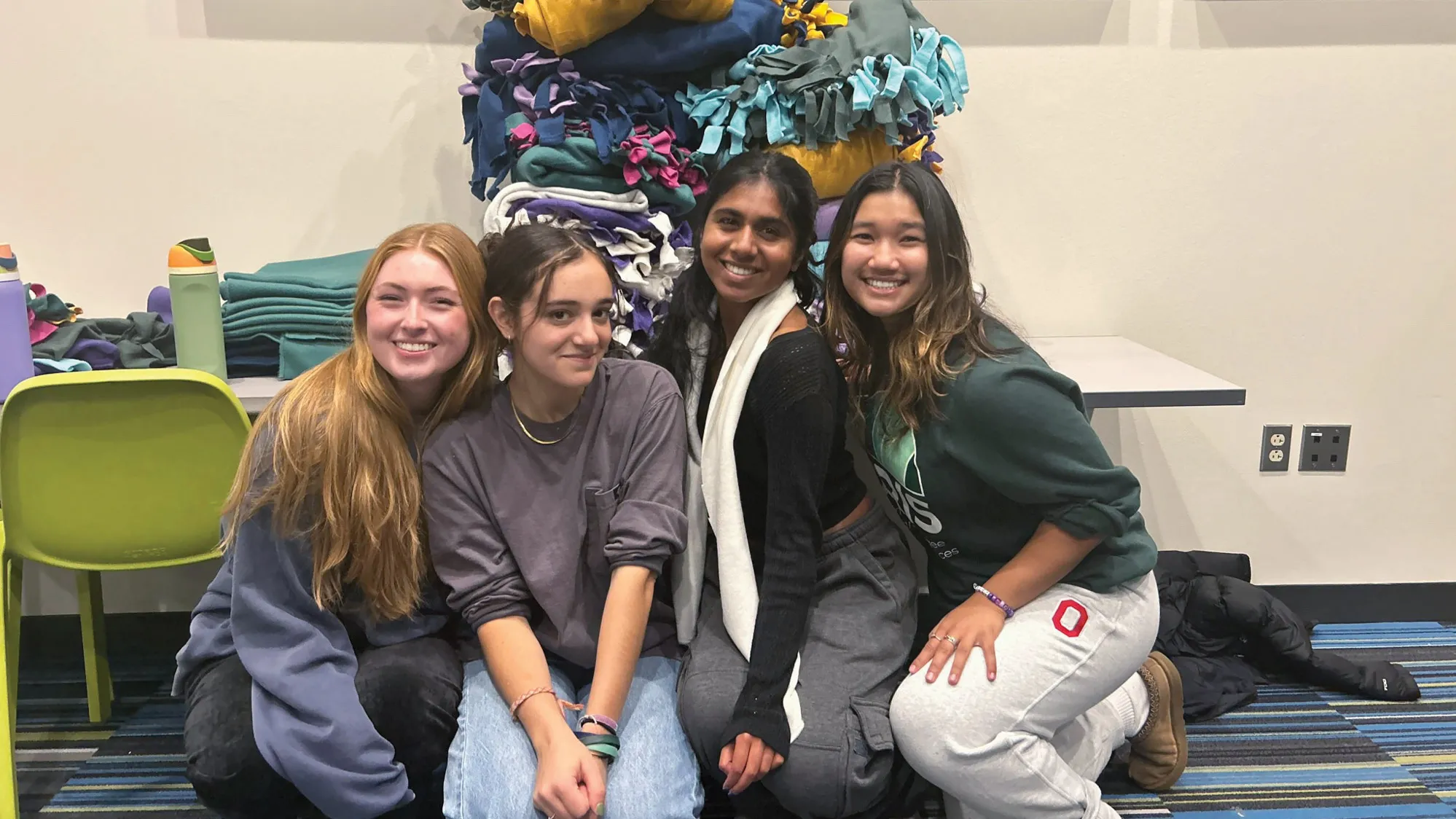 Crouching to pose in front of a table stacked high with fleece blankets are four female students smiling widely. The way they scooted close together speaks to the friends being very comfortable with one another. The blankets are double layered with wide fringe hand-knotted along each side.