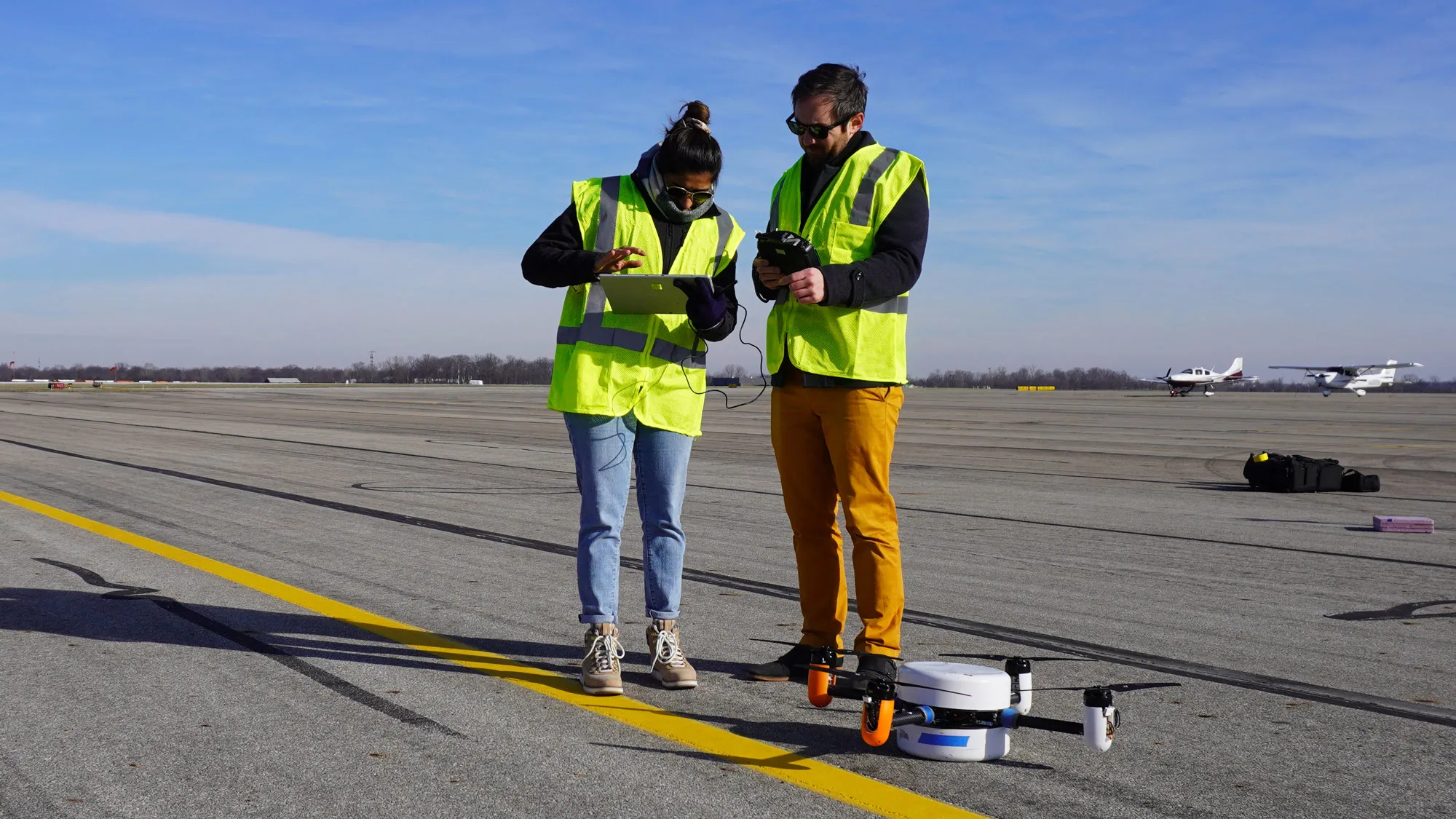 Two people, a taller white man and shorter woman working a large tablet computer, wear sunglasses and safety vests as they stand on what’s perhaps a runway at Ohio State Airport. A round drone with four propellers sits at their feet. 