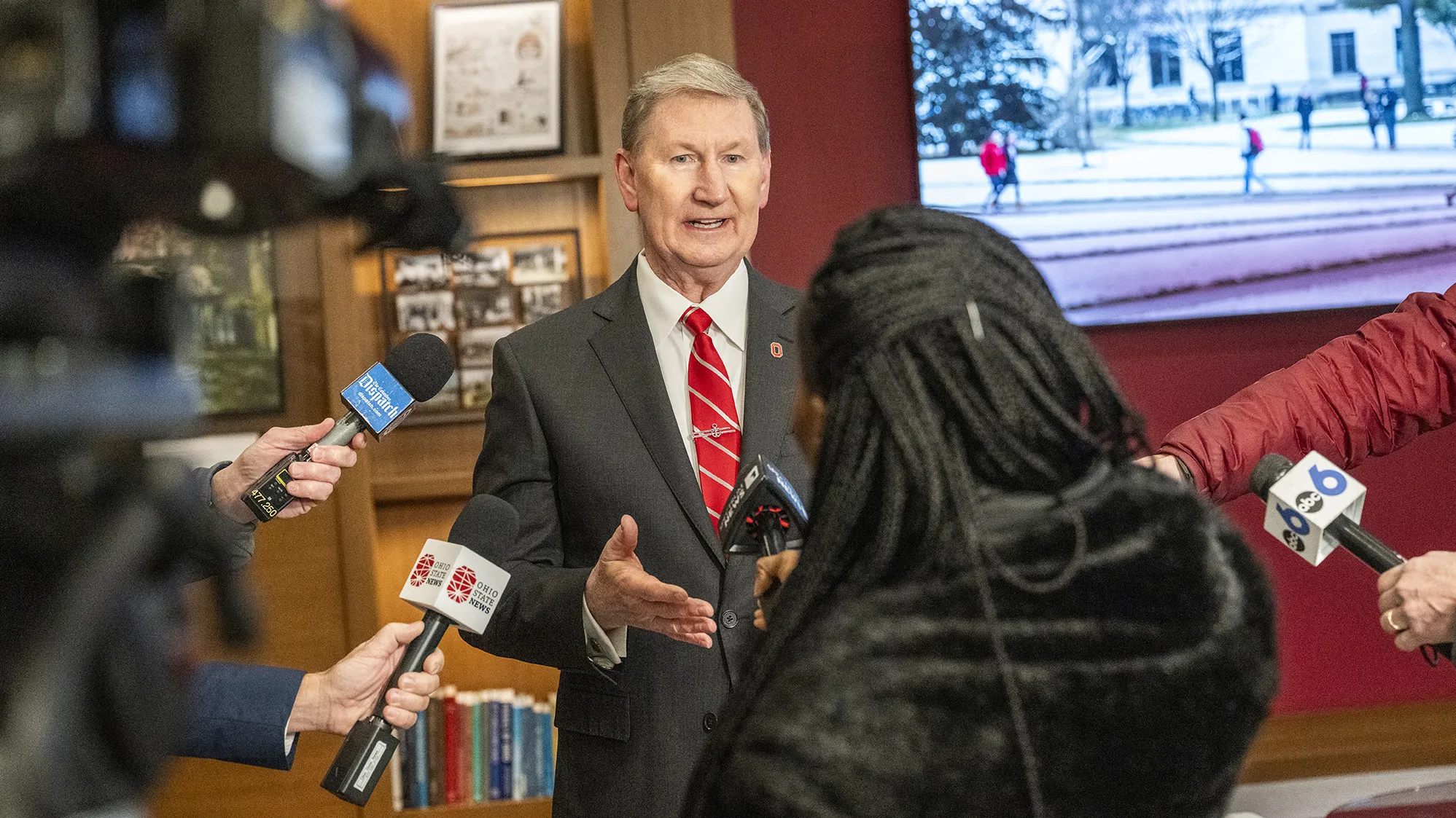 President Carter gestures as he answers questions from reporters circled around him. Most of the reporters don’t appear in the photo, but their outstretched arms holding microphones or cameras are aimed at him. The main reporter Carter is speaking to can be seen from behind. She has long hair in thin braids.