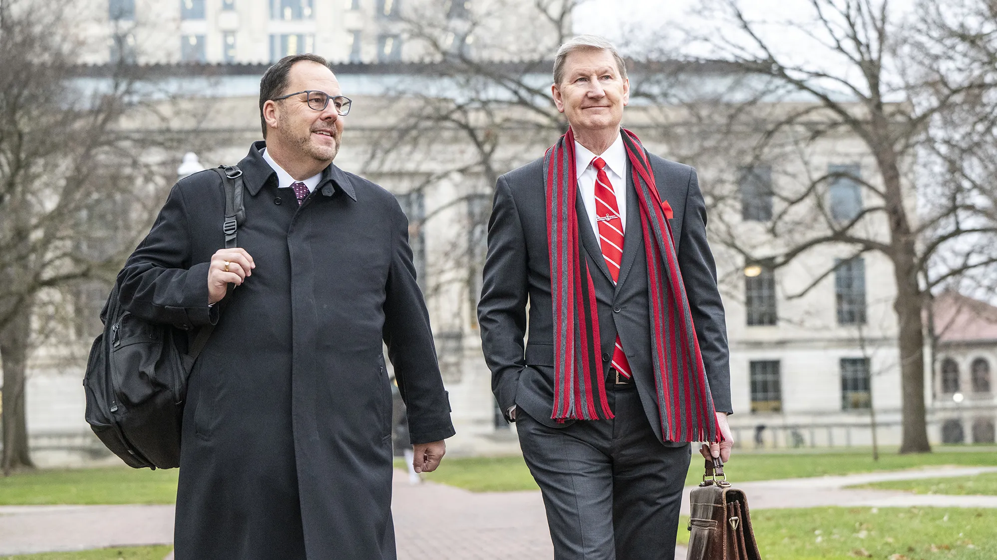 Ohio State President Ted Carter crosses the Oval with a confident but slight smile as we walks next to a university official. Carter wears a suit and long staff striped in Ohio State colors. JR Blackburn wears a black work coat and carries a backpack. In the background are stately academic buildings. The trees are bare but the grass is green