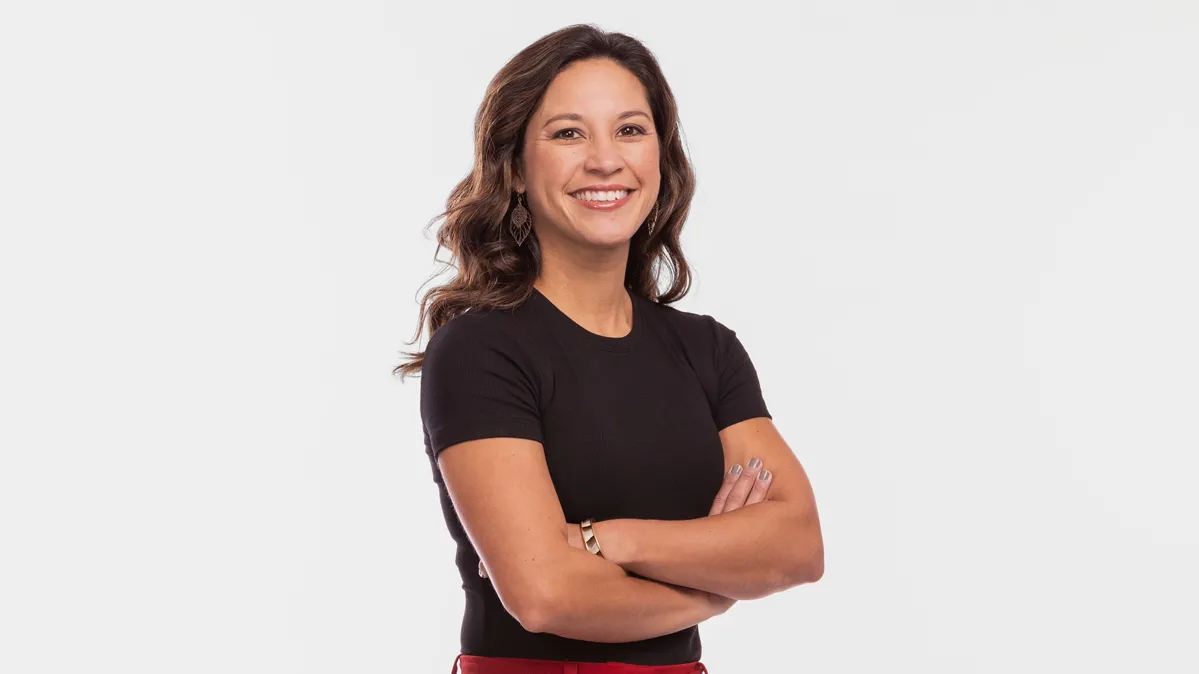 Stephanie Baxter, a white woman with long wavy hair, stands up straight, smiling with her arms crossed. She comes across as happy and confident as she poses in a fitted T-shirt, fashionable pants and heels.