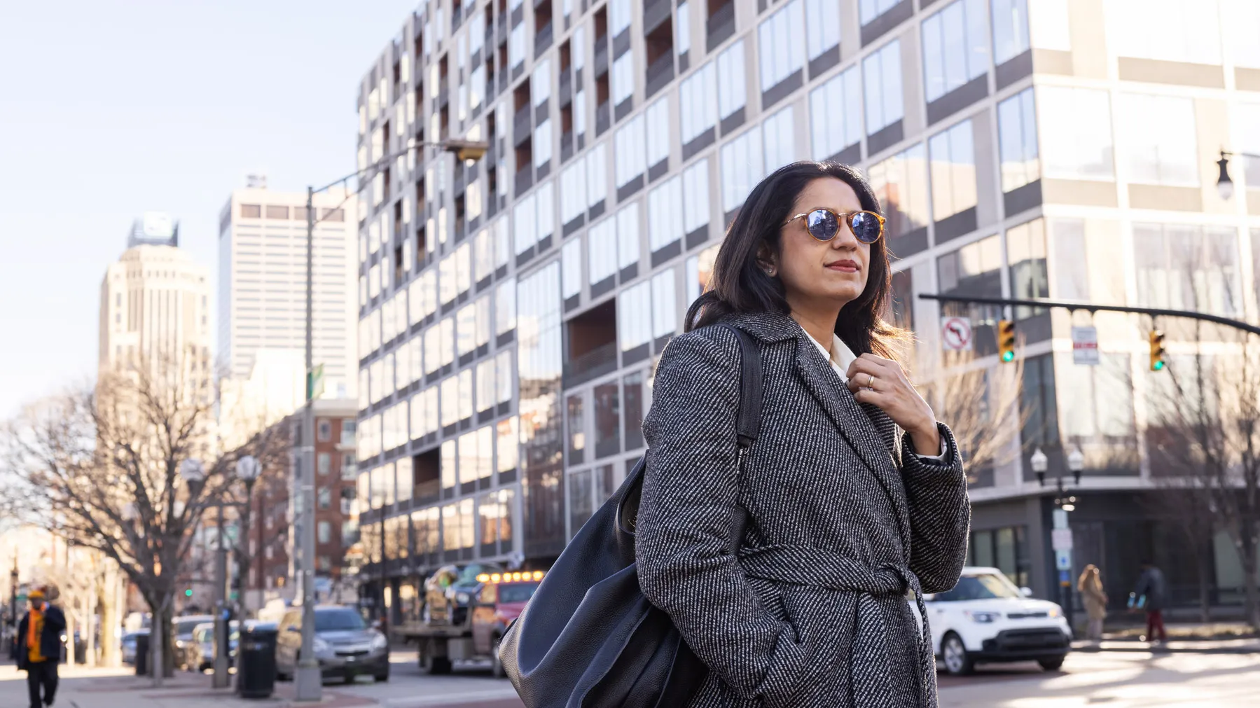 Waiting to cross a street in downtown Columbus, Bhakti Bania looks stylish and purposeful in round sunglasses and a chic coat. A skyscraper with shiny silver-tinted windows rises behind her.