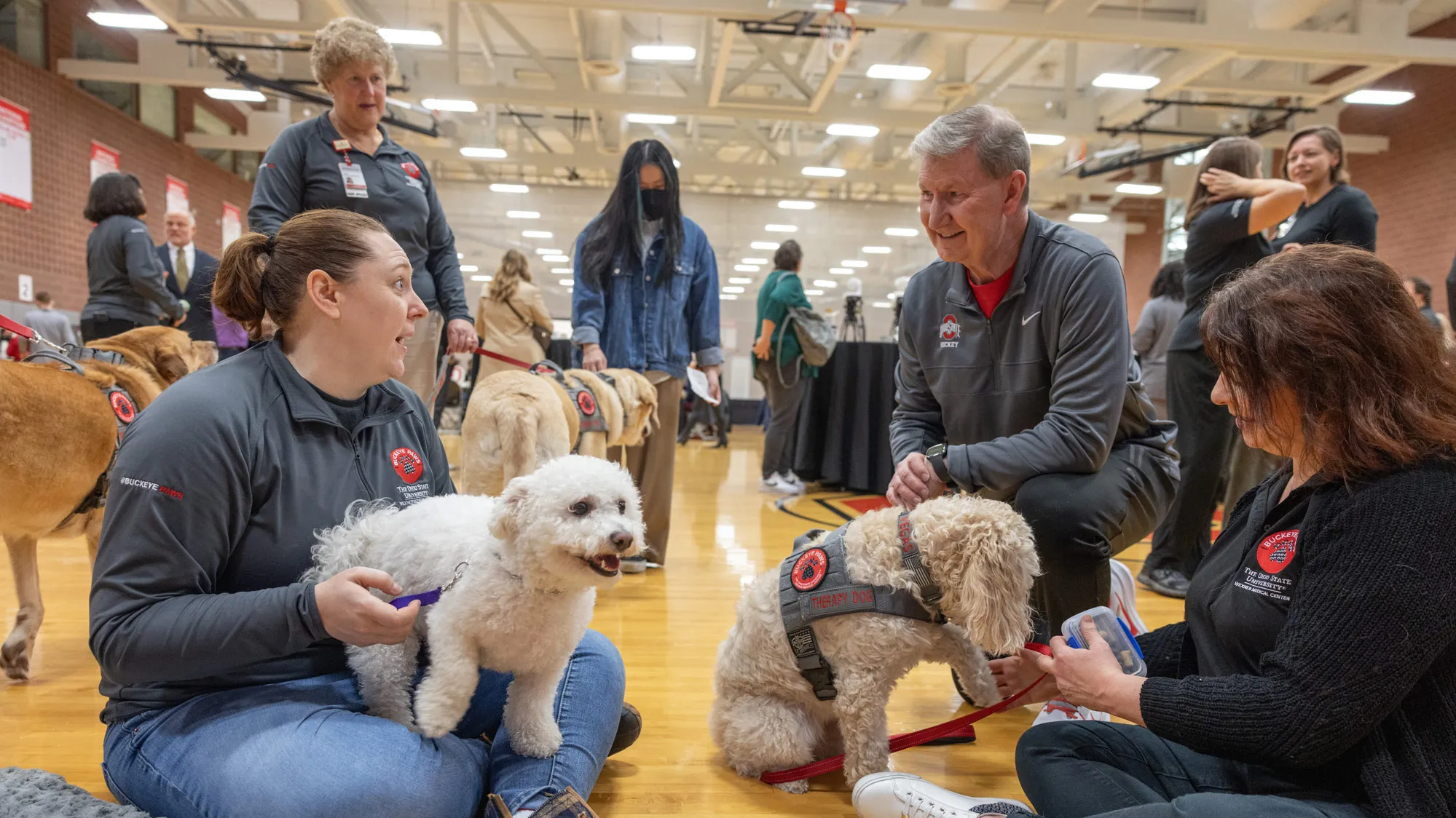 President Carter crouches on a gymnasium floor to talk with the owners of a pair of Buckeye Paws’ smaller dogs, both of which might be poodle mixes, judging by their fluffy, wavy fur. President Carter is chuckling as he makes eye contact with the woman talking, who is sitting on the floor and holding her dog on her lap. 