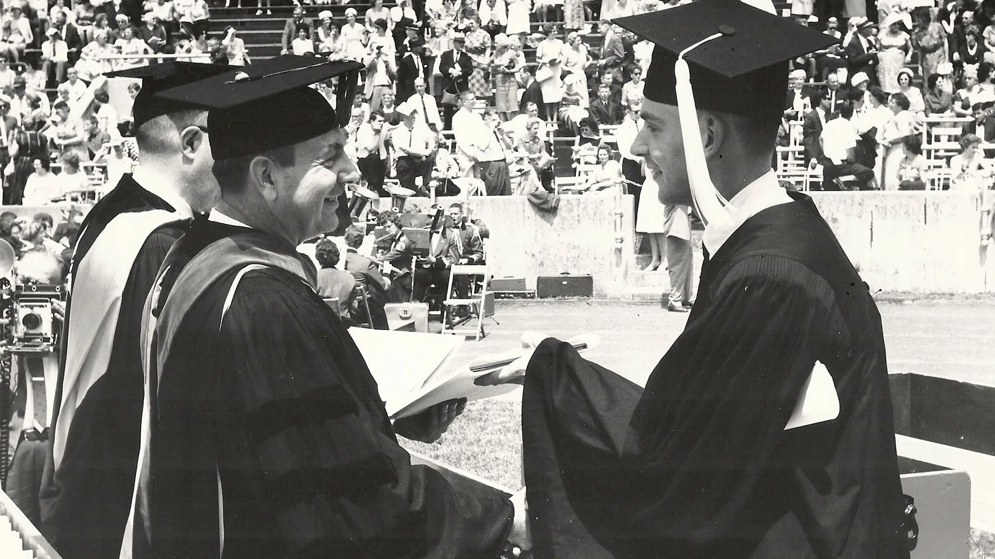 In a historical photo taken inside Ohio Stadium during a graduation commencement ceremony, a young man happily accepts his diploma from a pair of university leaders. All wear caps and gowns and smile. In the background, a crowd of spectators watches from the stands.