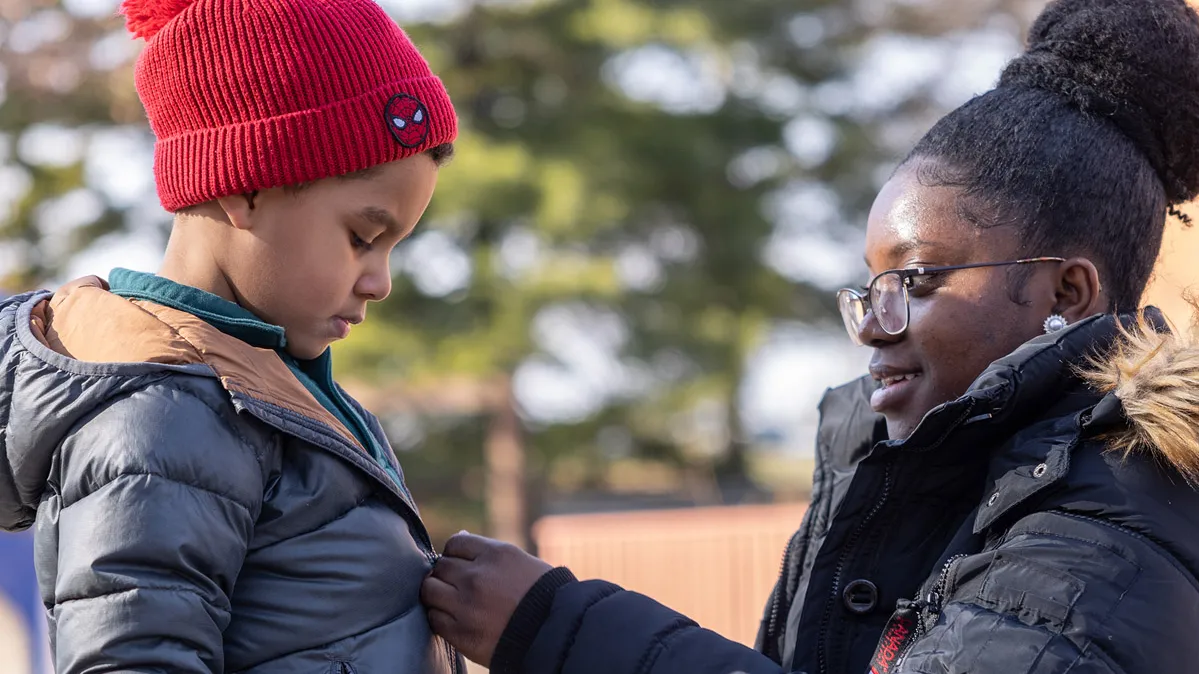 On an outdoor playground, a college student smiles as she crouches down to zip a child’s coat. She’s also wearing a heavy winter coat (along with glasses and her hair piled in a big bun on top of her head). The boy, wearing a stocking hat and gloves, watches her hands.