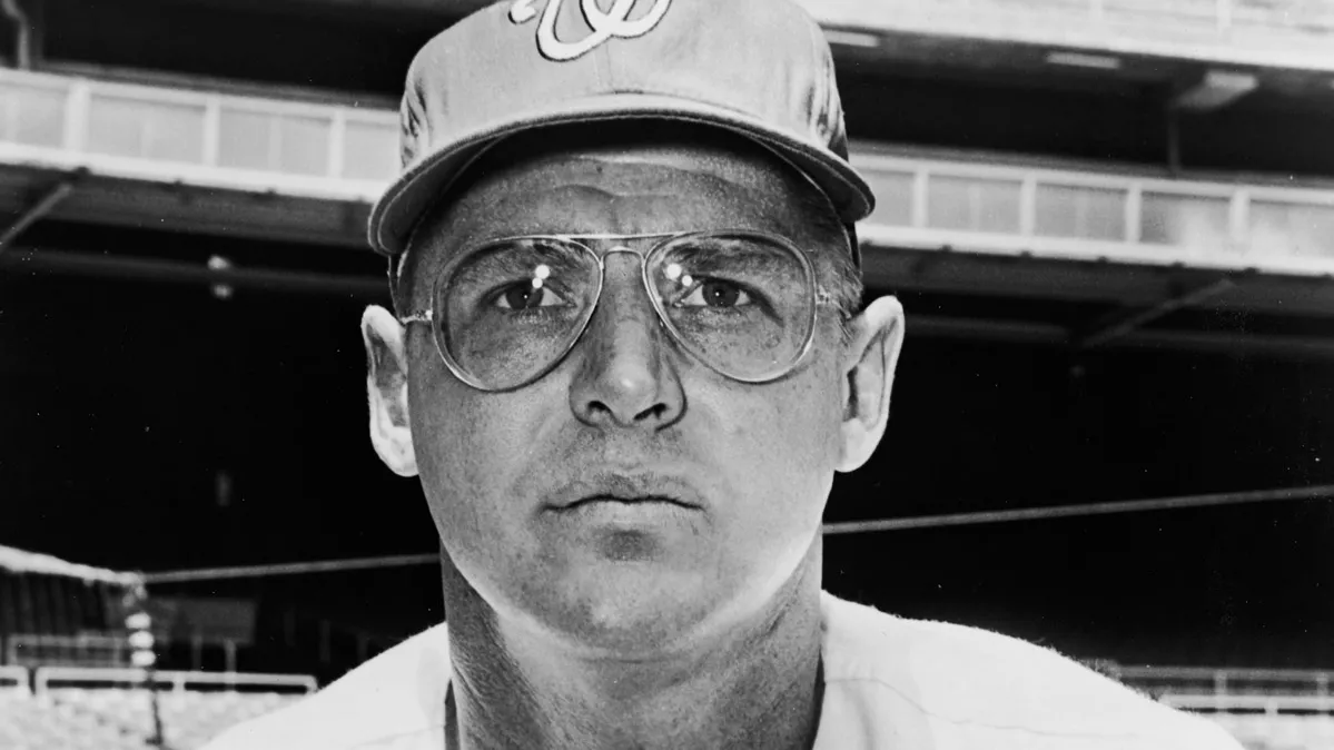In an older photo, a white man leans toward the camera while wearing his baseball uniform, glasses and a baseball cap. He looks focused and serious.