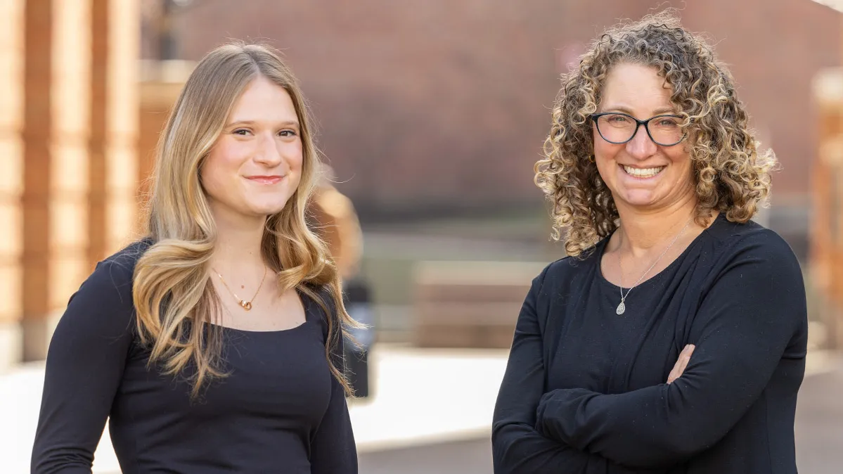 Two women stand in the courtyard of Ohio State’s Fisher College of Business. Both wear dark tops. The college student, on the left, has long blond hair and stands with one hand tucked in her pocket. The teacher, on the left, has curly shoulder length hair and stands with her arms crossed. Both smile as they pose.