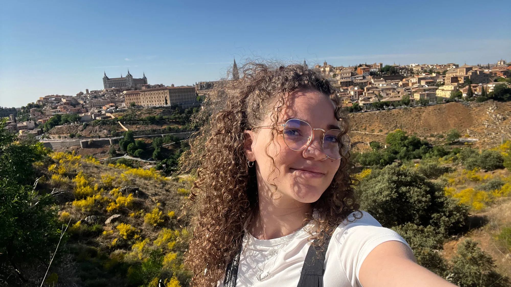 Ariana Winbush, a pretty young woman with long, curly hair smiles as she takes a selfie on a hill outside a town in Spain on a sunny day.