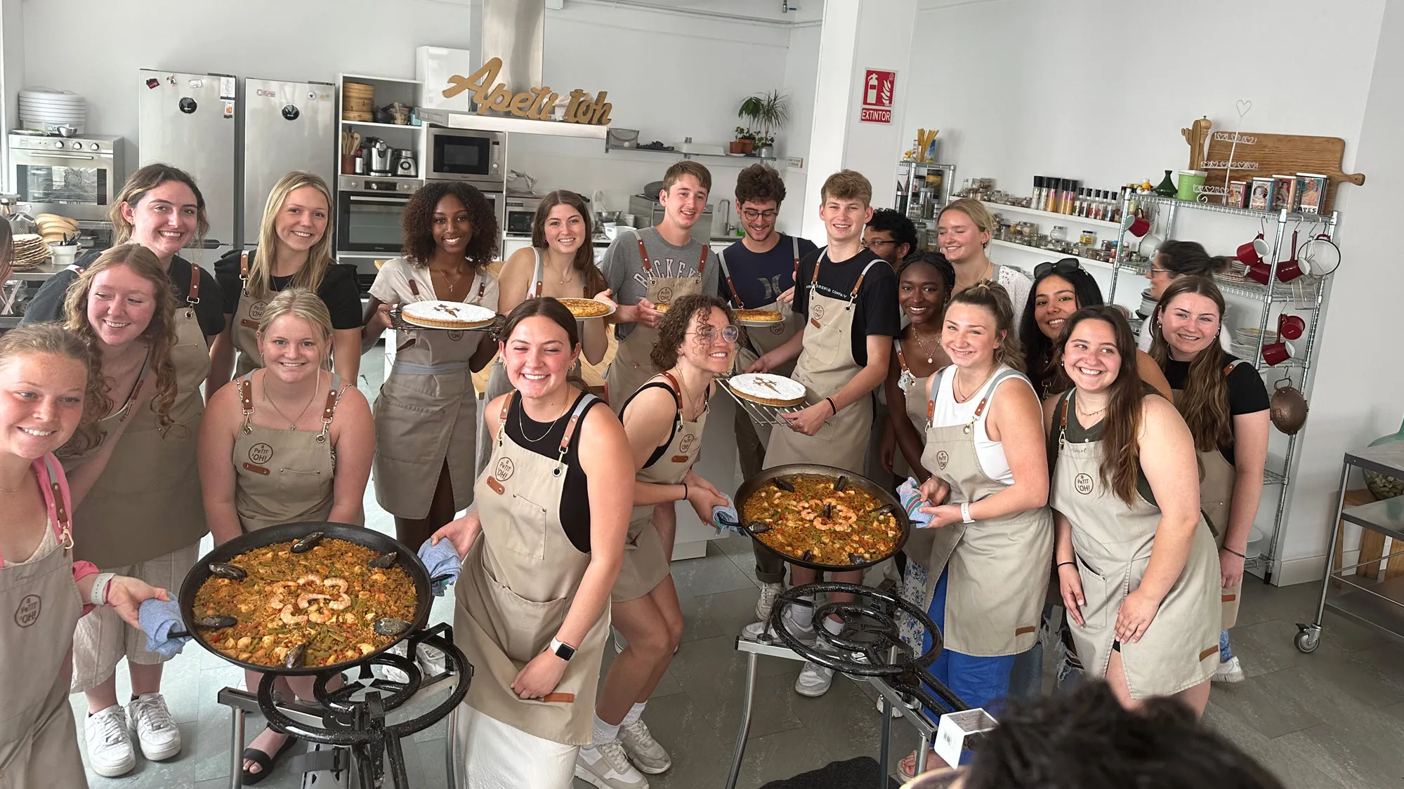 Twenty young people wearing matching aprons gather in a commercial kitchen around big pan-fulls of paella, a traditional Spanish dish.