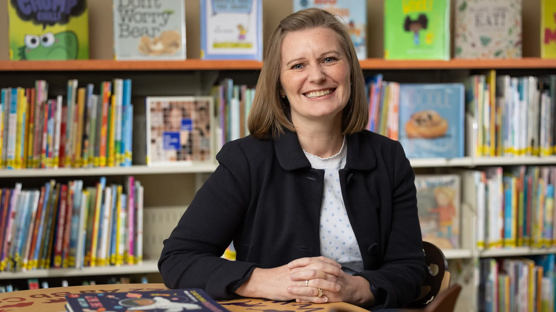 Shayne Piasta poses in a reading room surrounded by children’s books. Coming off as friendly and direct, she sits at a small table with her hands folded. She has a wide smile that crinkles the skin around her eyes and shoulder length hair.