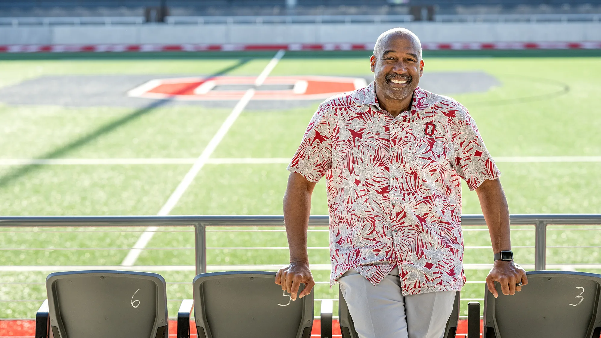 Wearing an Ohio State-colored tropical shirt—that looks perfect for retirement or vacation—Gene Smith grins as he leans on some front-row seats at Ohio Stadium.