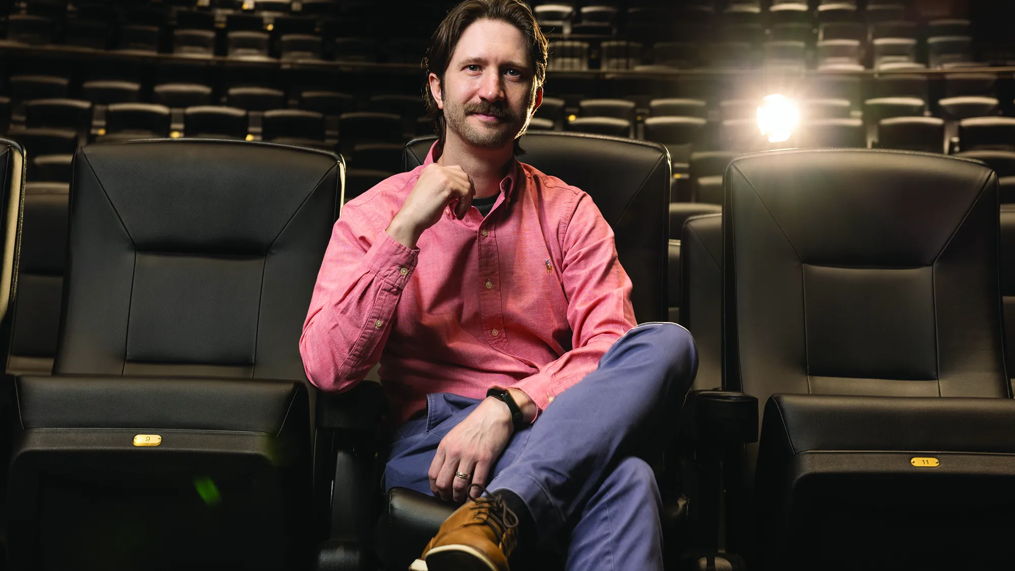 Sitting in a theater with leather seats, Matthew Grizzard, a bearded white man wearing khakis and a button down, sits relaxed with one leg crossed over the other and his hands seeming as if he’s focusing as he listens. He’s smiling with his eyes more than his mouth.