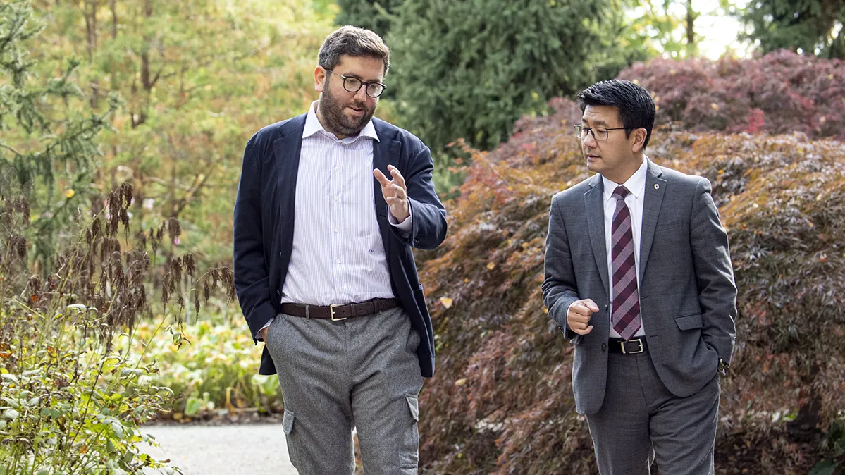 Walking on a gravel path through an area with lush plants and trees, Jeffrey Schottenstein, a bearded white man wearing a cardigan, button-up and glasses, gestures as he speaks with Dr. K Luan Phan, a man of Asian descent wearing a suit and tie. He’s listening intently and the men’s heads are canted toward each other as they look forward