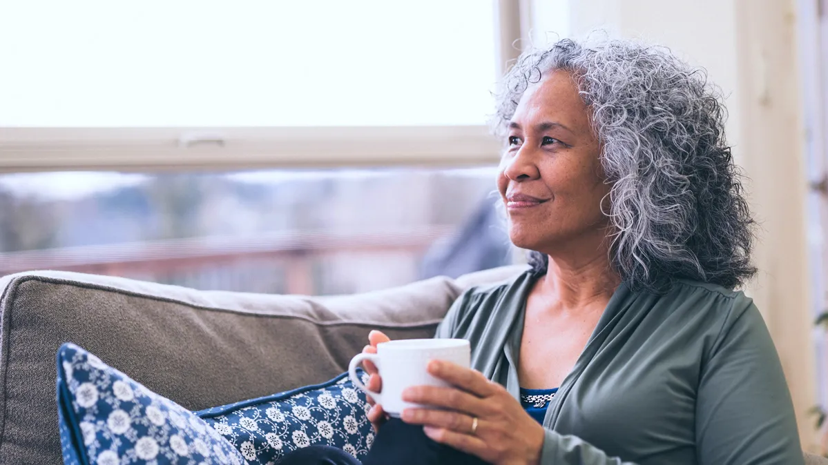 A woman sits with knees to her body near a window and holding a hot beverage. She appears contemplative as she looks into the distance