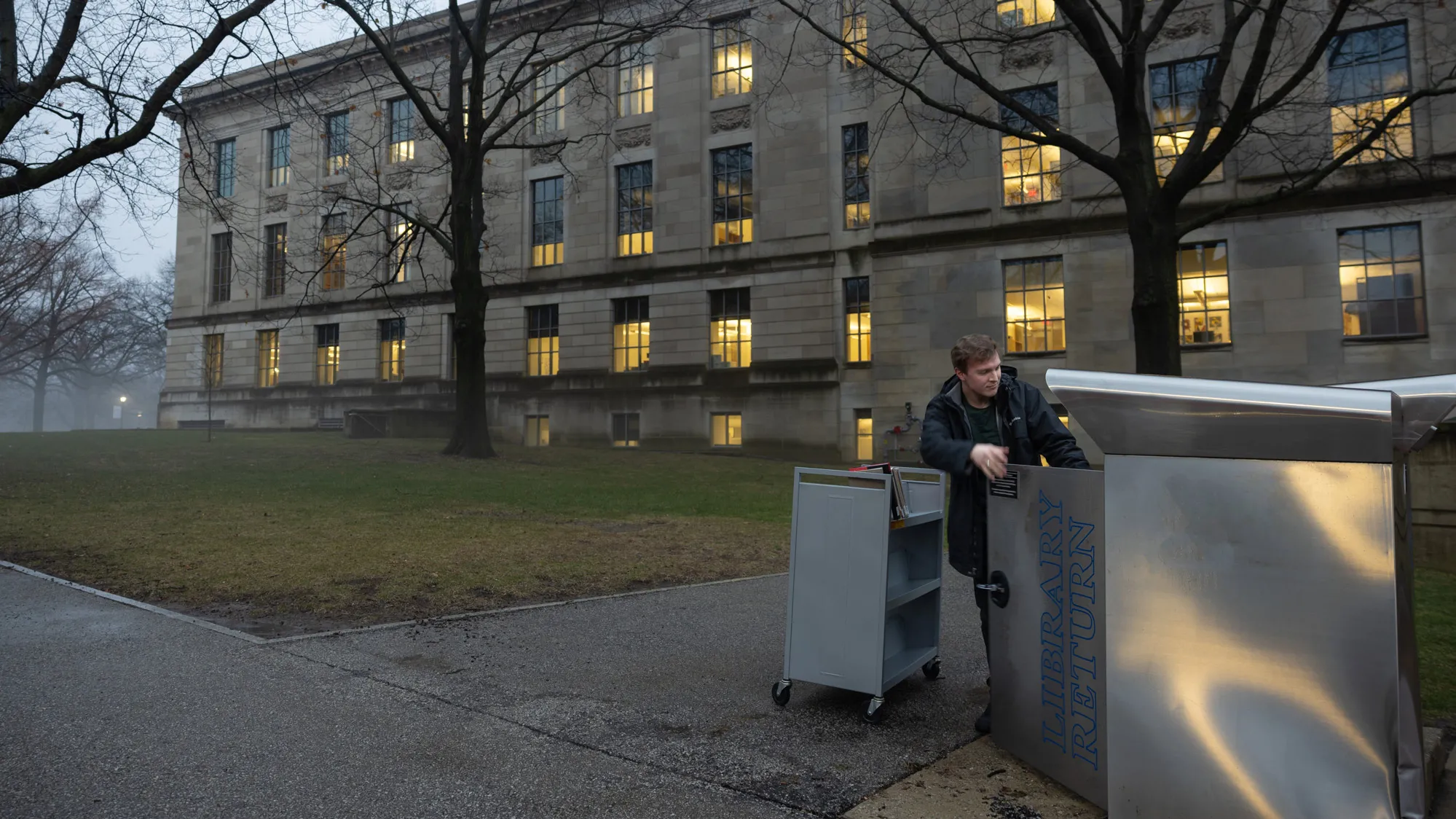The light outside the library is dim, indicating the sun has begun to set. Warm lights shine from the windows of the north face of the library. A young white man in a winter coat has rolled a cart to a large metal bin—it’s almost as tall as he is—to collect the books others have returned there. 