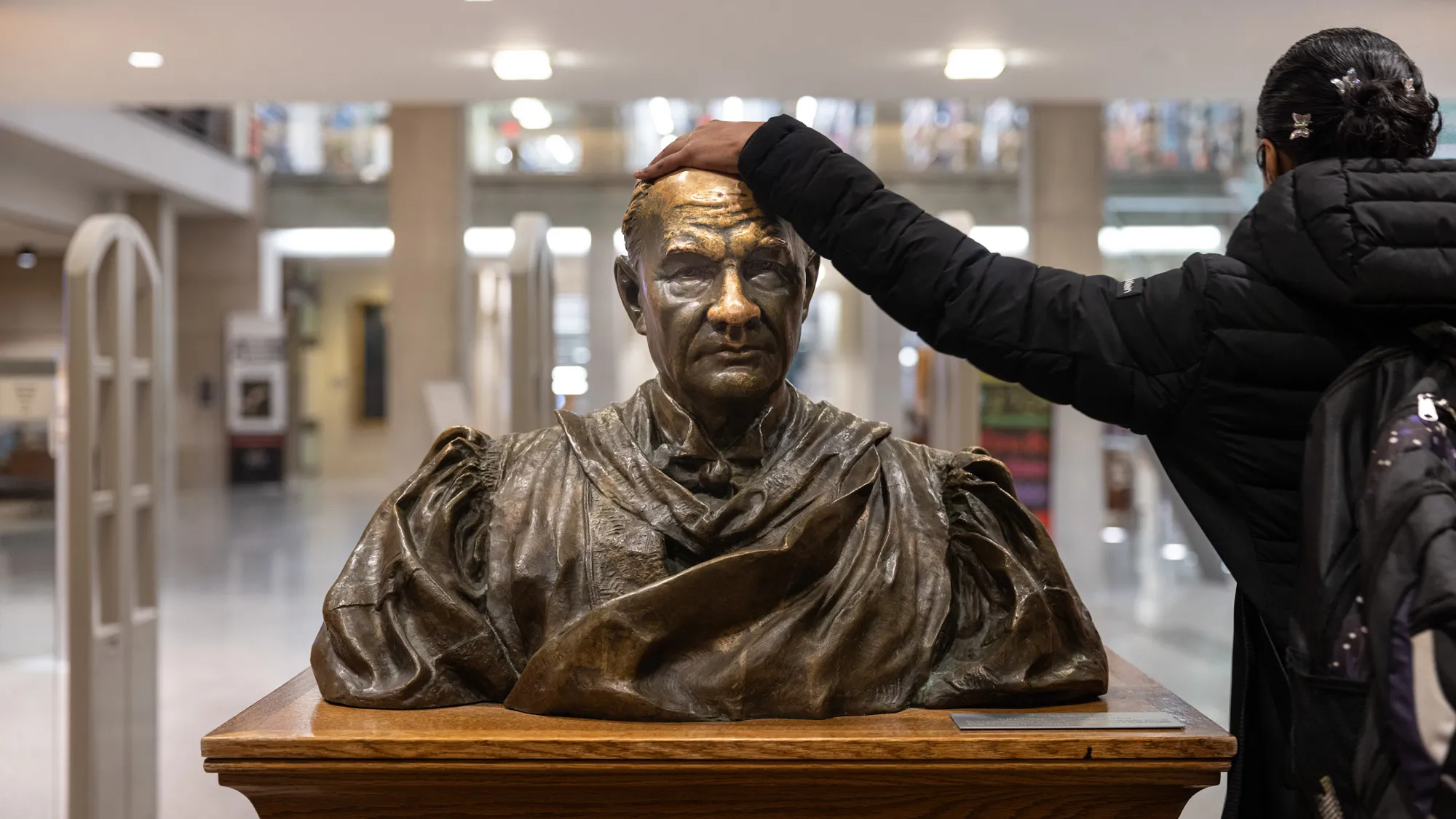 A bronze statue on a wooden pedestal shows a man’s face and chest, shoulders and top of biceps. He looks straight ahead with a serious, perhaps even studious, expression. His forehead and nose—where the statue gets touched a lot—are brighter than other parts, creating dramatic differences, such as on the wrinkles of his forehead. A student seen from behind pats the statue’s head as she walks in. She wears a long winter coat, a backpack slipped over both shoulders and her black hair pulled into a bun.  
