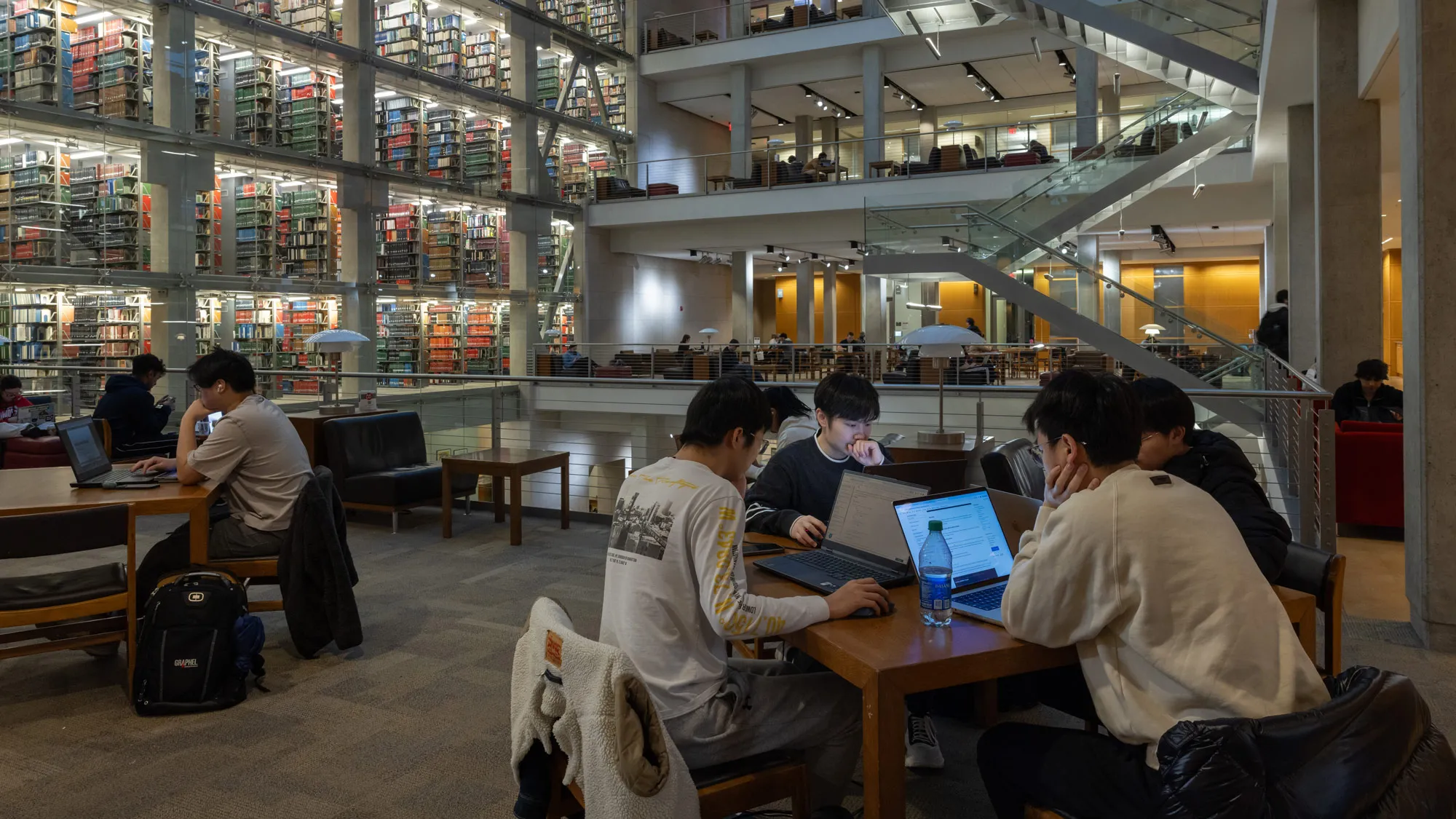 With the light inside the library dimmed because it’s dark outside (the library’s huge skylight provides much natural lighting), four young Asian men sit at a square table, each engrossed in his own laptop.  