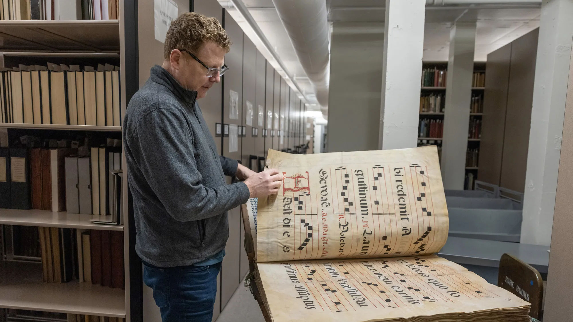 A white man with wavy hair and thick-rimmed glasses pages through a volume that’s as tall as his torso. The content is music notes with words in another language. 