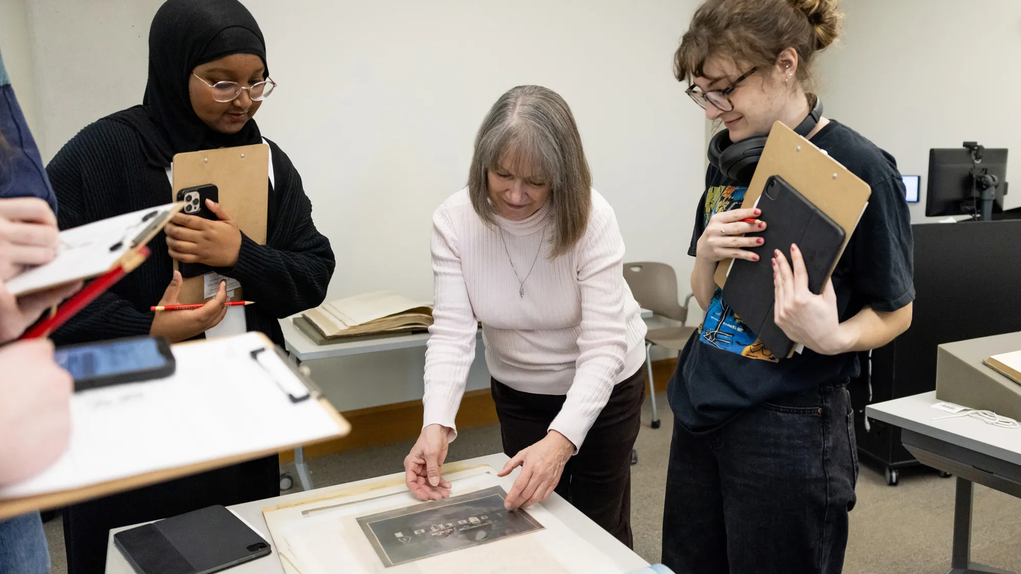A librarian, an older white woman with neat, shoulder length hair, leans forward to touch a historical photo lying on an open book as students stand around her her holding clipboards and devices. Two can be seen: Right of the librarian, a young white woman has thick rimmed glasses. Left of the librarian, a young Black woman wears a hijab and thin, wire-framed glasses. Both students seem intrigued. 