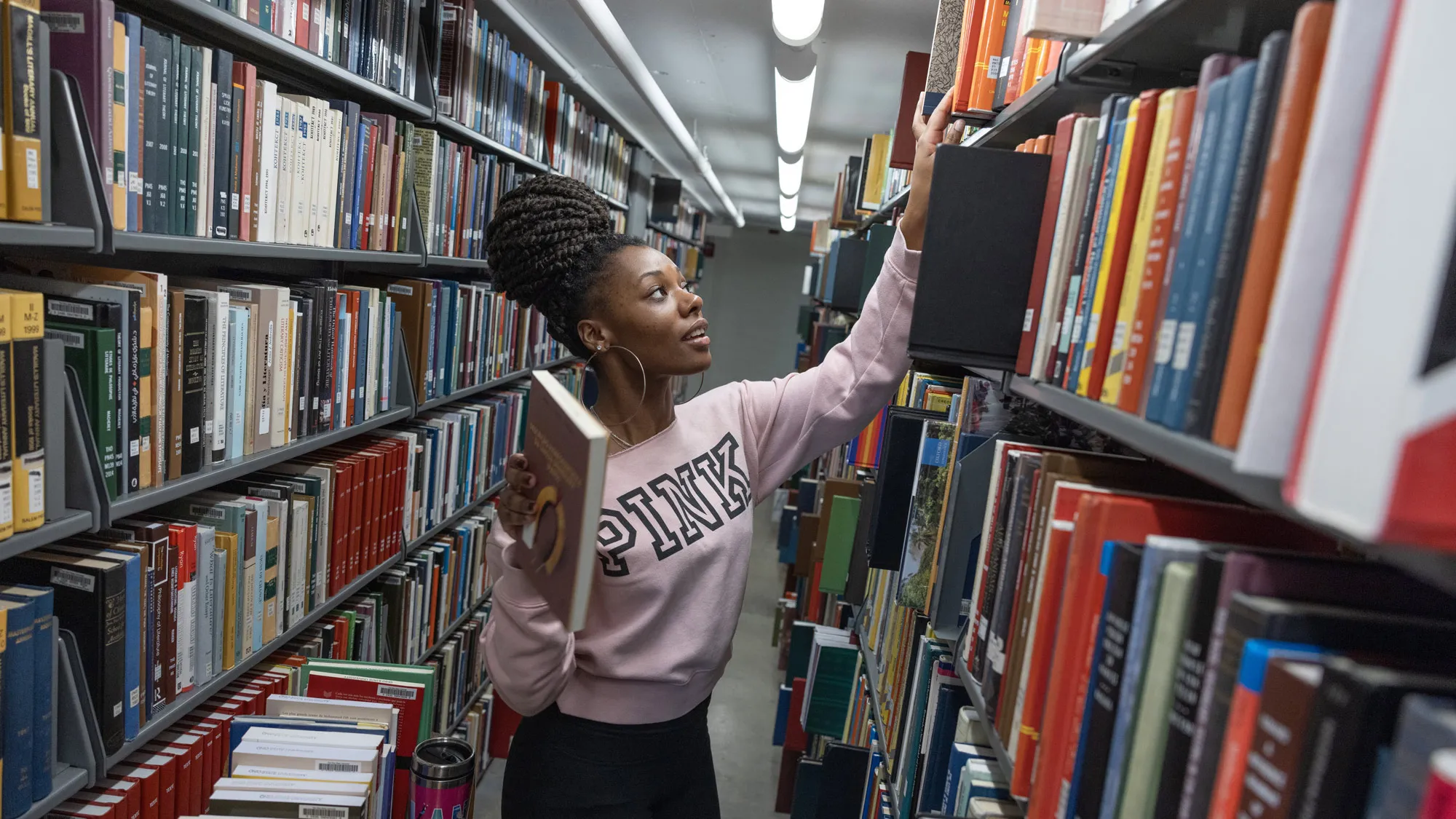 Deep in some of the stacks, a young Black woman, with her hair twisted into a tall stylish bun on top of her head, reaches above her head to shelf a large book. She holds another in her other hand and, though frozen in this moment by the photo, appears graceful in doing her work. Her sweatshirt says “pink” in big capital letters. 