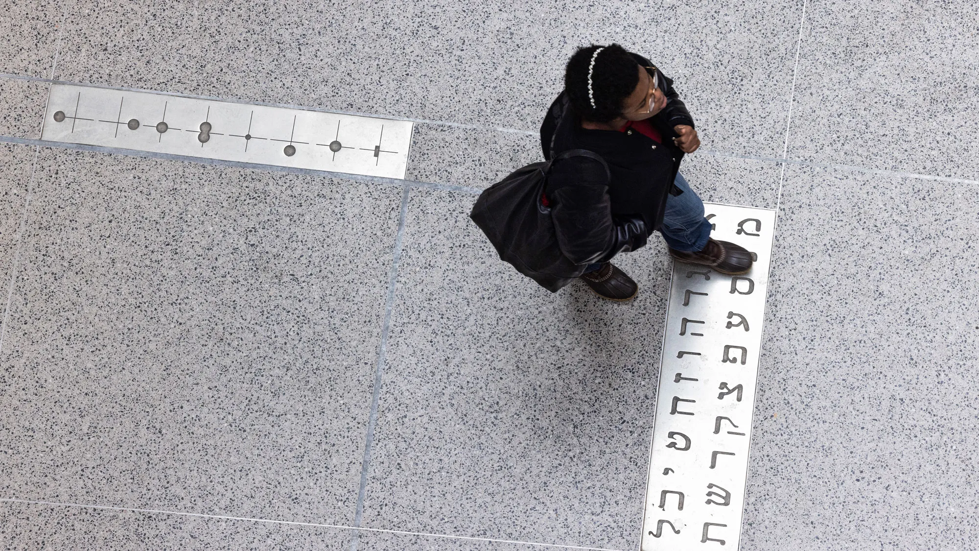 Photographed from directly above, a Black student crossed the library’s lobby floor. It is composed of large slabs of a material that appears to be granite or marble speckled in tones of the same gray shade. Inset in two pieces of matte metal—one under the student’s feet and another behind the student—are other types of language. 