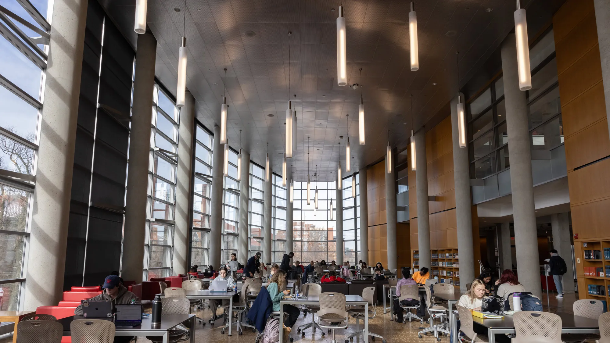 A crowded room in the library has modern, tall and slim lights—at least 20 of them—hanging from the ceiling; walls made of glass paneled windows or sleek wood and columns; and a floor covered in embossed text. The letters are hand-sized. More students than can be counted study at sleek tables in plastic rolling chairs. 