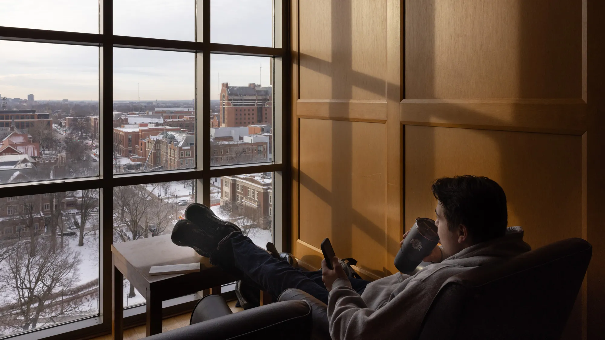 In a cozy scene, a young man kicks back in an arm chair, his legs propped on a coffee table, while holding his cellphone and sipping from a travel mug. Through the window in front of him, he looks down on several blocks of a snow-covered campus. Red brick buildings stand out and morning light creates a modular shadow pattern on the wooden wall beside him.