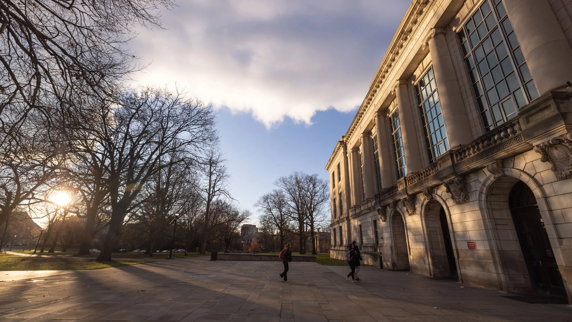 In the morning light on a cold day (indicated by leaf-less trees and the distant people walking toward the library while wearing winter coats), clouds against a blue sky cast shadows on the columned front of the library. Multi-paned windows reflect the blue of the sky above a few arched entrances. 