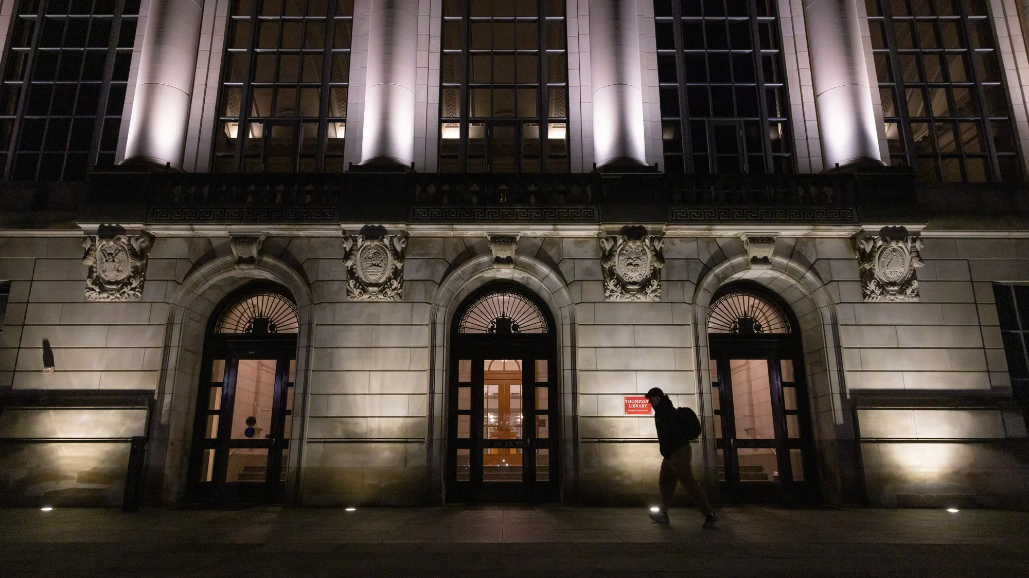 At night, a student passes by on the Oval side of the library. Only the silhouette of the student can be seen. Lights shining on the building highlight the stately architecture. 