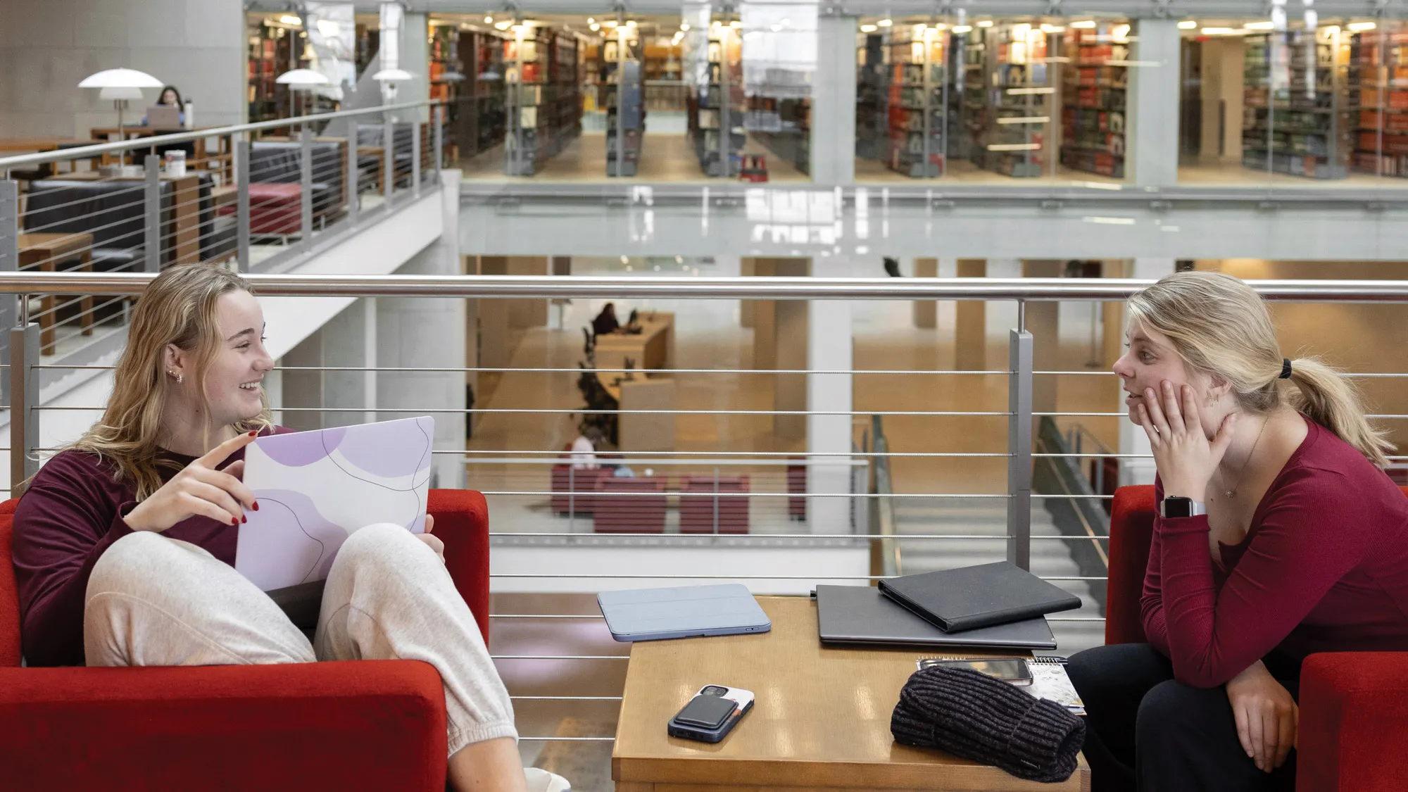 Sitting in armchairs in an atrium above the first floor facing each other, two young blond women smile and chat. Their body language says they’re good friends, and a closed laptop and face-down cellphone sit on a small knee-high table between them. Four floors of the glass-fronted stacks, revealing scores of book-filled shelves, make up the photo’s background. The scene is modern and brightly lit, showing the optimistic feeling of Thompson Library. 