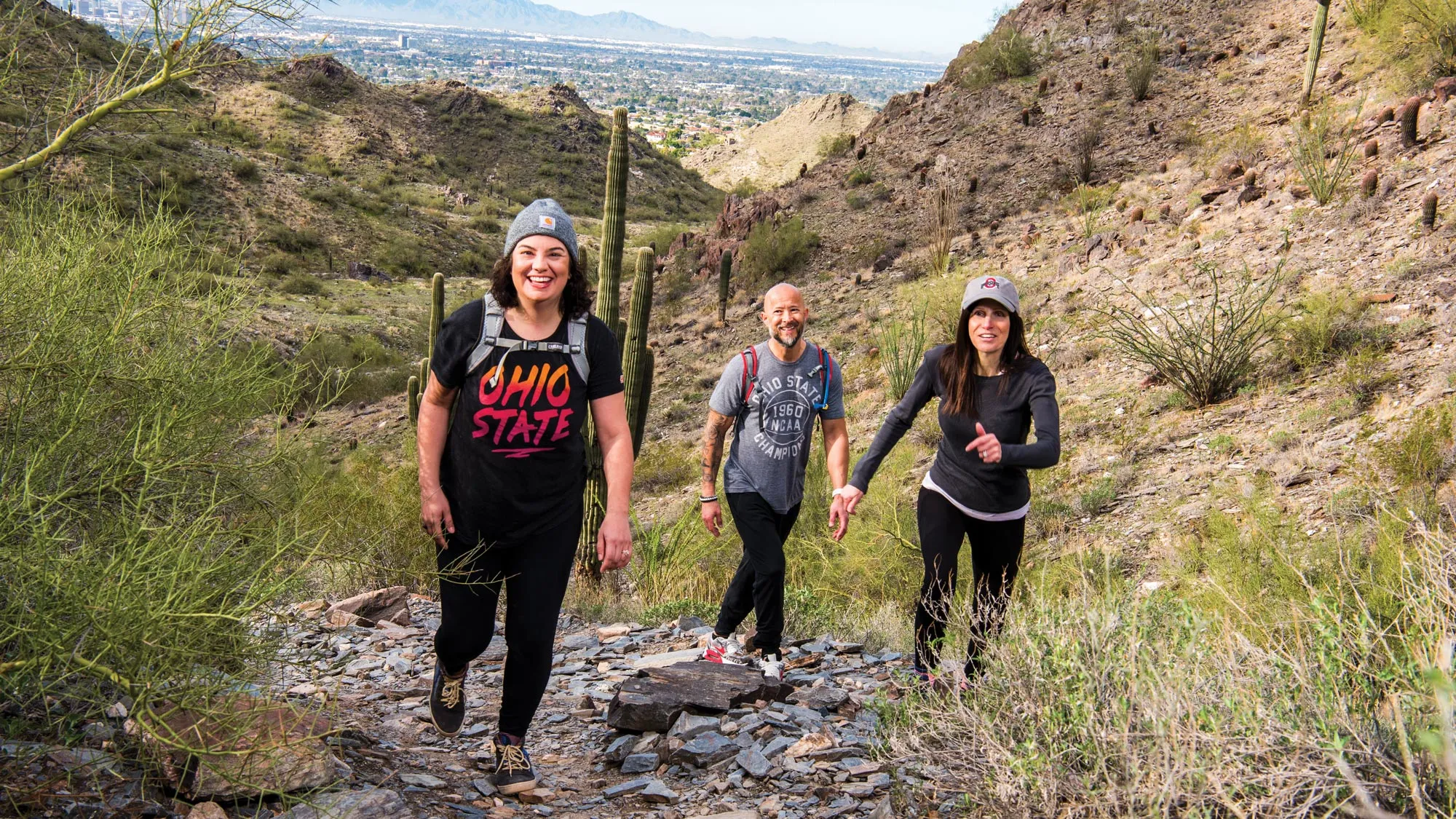 Climbing up a path littered with flat rocks, three Ohio State alumni grin as they look toward the photographer ahead of them. Around them the landscape is nothing but hills, brush, dirt and plants, including the occasional cactus.