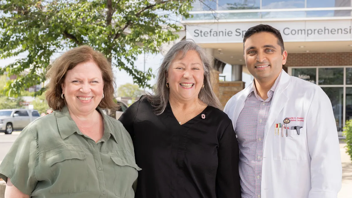 Three people pose for this photo in front of the entrance to the Stefanie Spielman Comprehensive Breast Center, part of The Ohio State University Comprehensive Cancer Center – Arthur G. James Cancer Hospital and Richard J. Solove Research Institute. The three are Molly Ranz Calhoun and the doctors who treated her cancer. They stand closely as they pose, seeming like they have a good relationship, and all smile as they squint a bit in the bright sunshine.