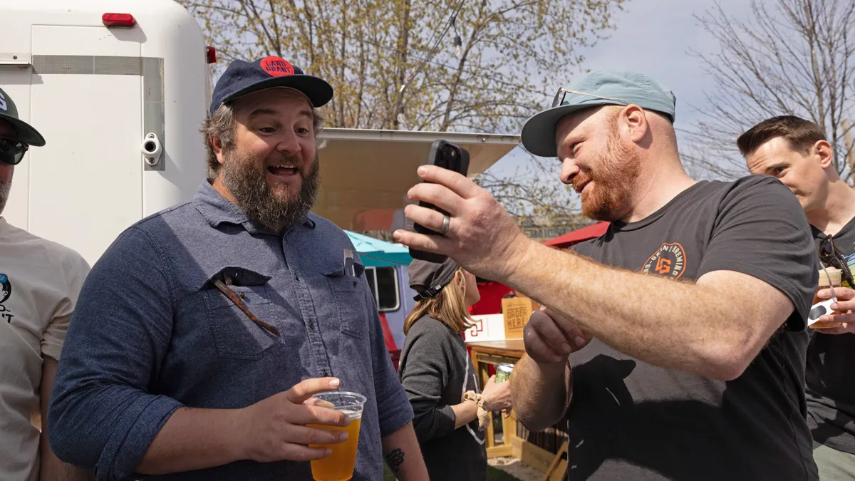 Adam Banner chuckles as he shows friend and work partner Walt Keys something on his cellphone. Walt’s expression says he is surprised and delighted. Both men are white with beards, wear ball caps and have sunglasses somewhere on them—for Adam, on his hat; for Walt tucked into the pocket of his button-up. Walt holds a beer in a plastic cup and they’re among many people as it’s their eclipse-day event.