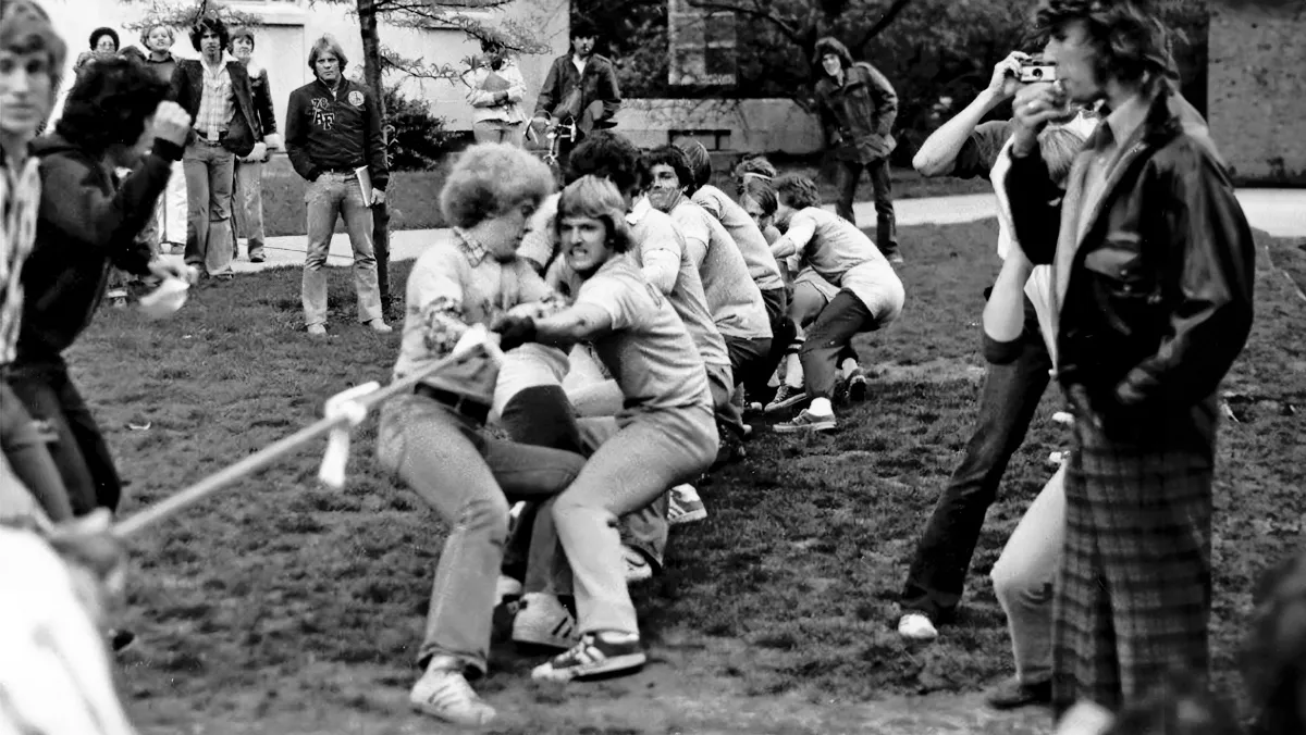 A team of young men, at least nine people strong, grimace as they pull with all of their might on a rope in a tug of war contest. The opposing team is out of the frame. The photo itself is black and white, as it was taken in the 1970s. The people’s clothes and hair styles makes that very apparent.