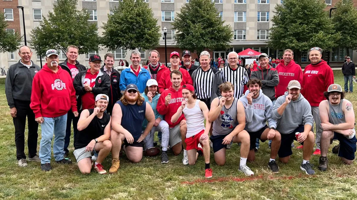 Two lines of people—alumni tug-of-war referees and coordinators and student participants—pose for a photo. The students—the team that won the competition—all bite their medals in a joking manner, as if testing to make sure the metal is real gold.