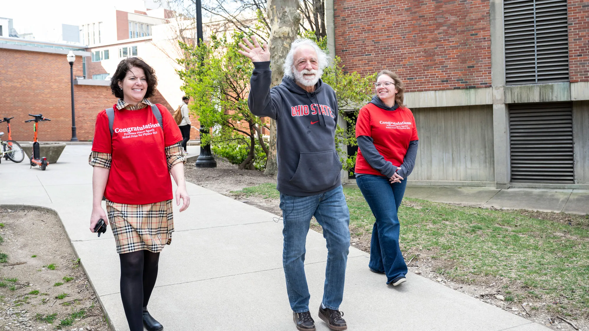 Agostini waves while walking toward a crowd of people. He is flanked by two white women who are university employees wearing Ohio State T-shirts that say “Congratulations.” They’re both grinning, and all three seem to be enjoying the cheers of the crowd, people who are not shown in this photo. Agostini wears an Ohio State sweatshirt. 