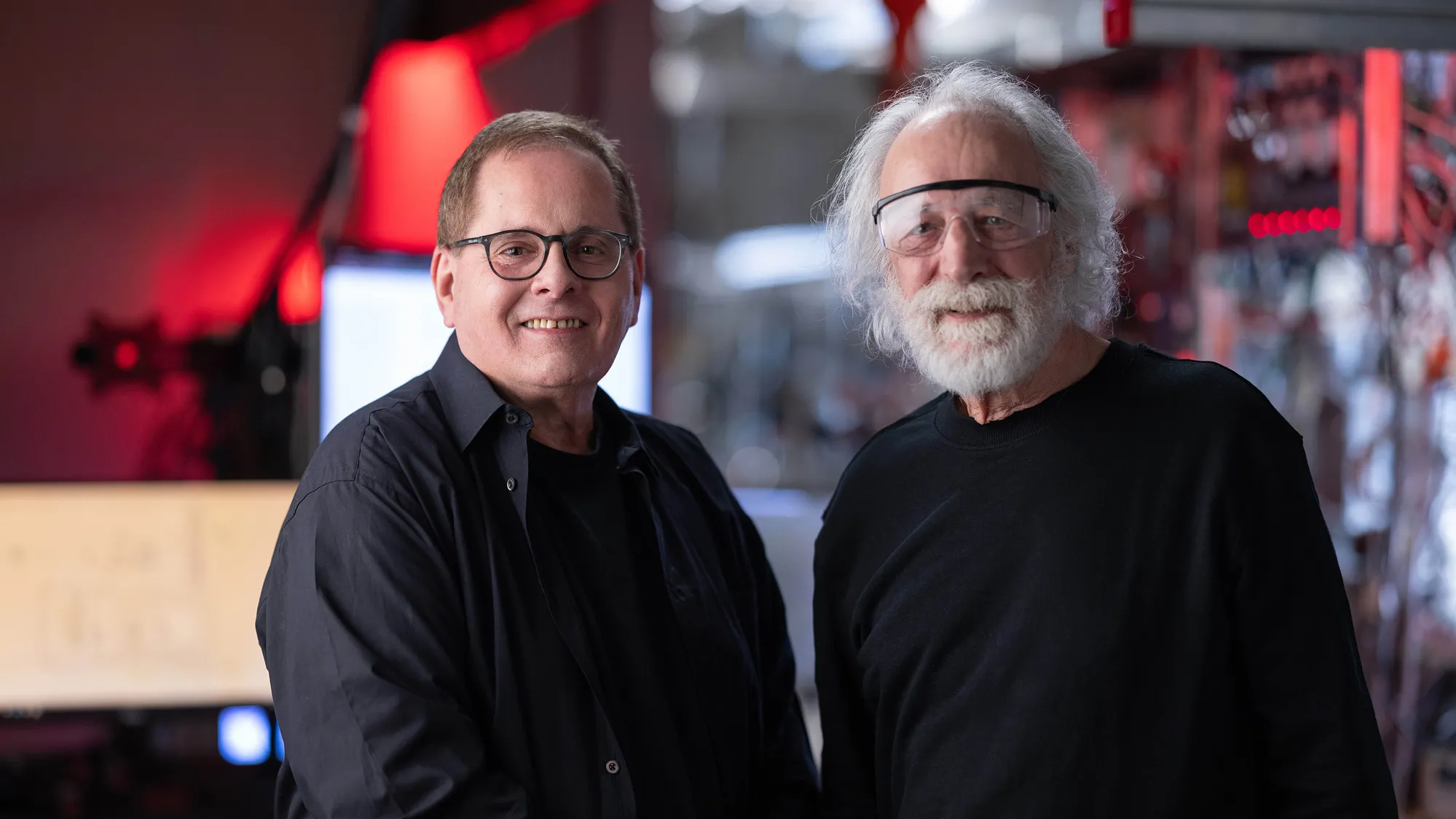 Lou DiMauro and Pierre Agostini smile as they pose for a portrait. Pierre has flowing white hair and a beard and wears safety goggles. Lou has short brown hair and glasses. Both are white men who radiate friendliness while dressed casually. 