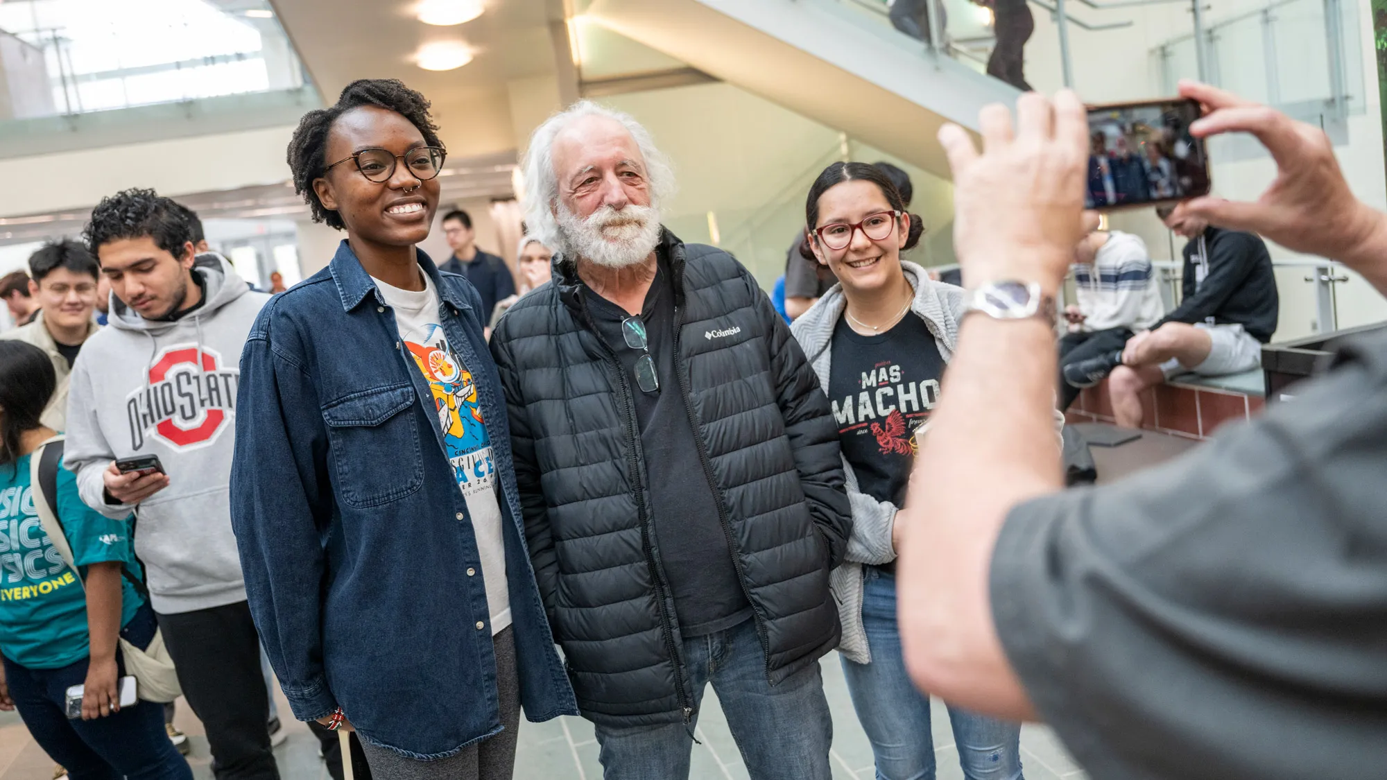 In a black T-shirt and insulated jacket, Agostini poses with two students for someone taking a cellphone photo. On his right, the black woman is taller than he is; on his other side, the woman (whose race isn’t clear) is as tall as his nose, so it looks like they’re standing in order of height. Both students wear glasses, T-shirts and smiles, and a line of students behind them wait their turn to meet Agostini. 