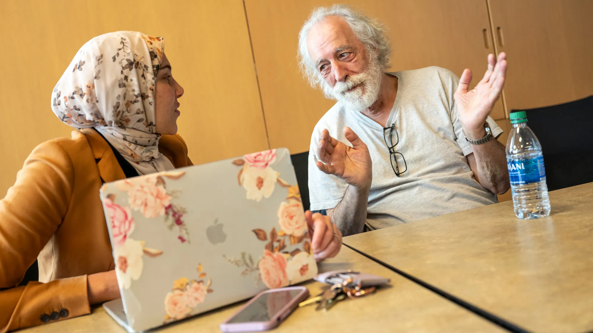 A student wearing a floral-print headscarf and suit jacket listens as Agostini, in a T-shirt, looks her in the eyes as he answers her strong-field physics question. She’s a member of his research group and is originally from the Middle East.  On the conference table in front of them are his bottle of water and her laptop, cellphone and keys. 