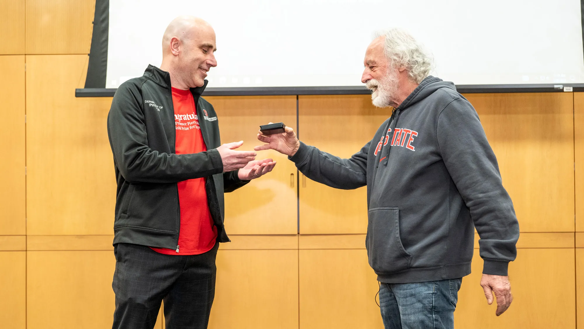 Standing at the front of a wood-paneled conference room, Agostini (wearing his favorite Ohio State sweatshirt) hands a palm-sized, shallow box to Michael Poirer, the head of the Physics Department. Poirer, a white man with a shaved head, reaches to accept it. The inside can’t be seen, but it holds a replica of Agostini’s Nobel Prize medal. 