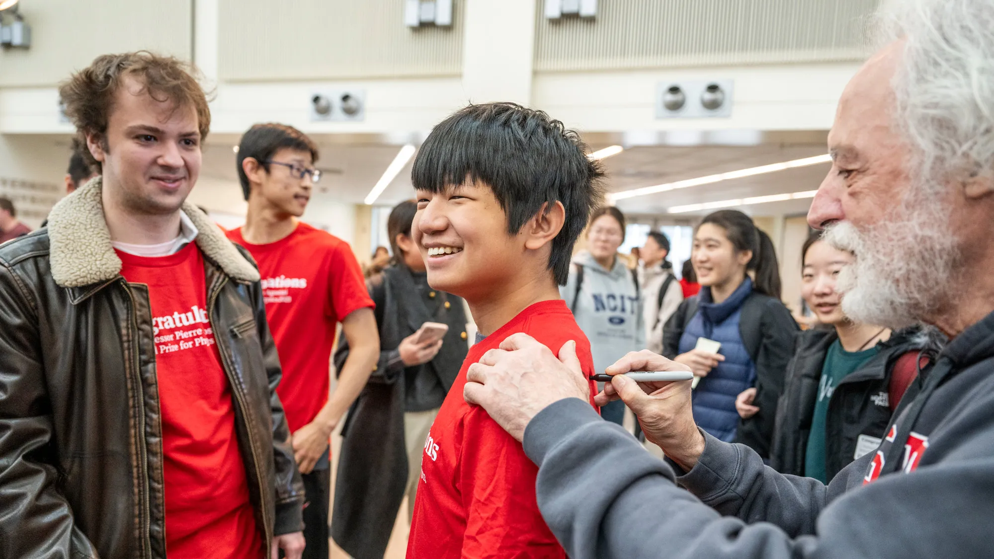 Agostini uses a Sharpie to sign the back of a T-shirt worn by a student of Asian descent. The young man grins and others around them smile as they watch. 