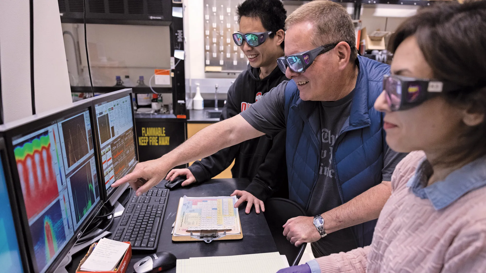 Three people—DiMauro and two of his students, a man originally from China and a woman originally from the Middle East—look at a screen displaying colors and graphs that reflect how electrons moved in their experiments. As DiMauro points at one, they all smile. Since they are inside their lab, which uses lasers, they all wear safety goggles. 