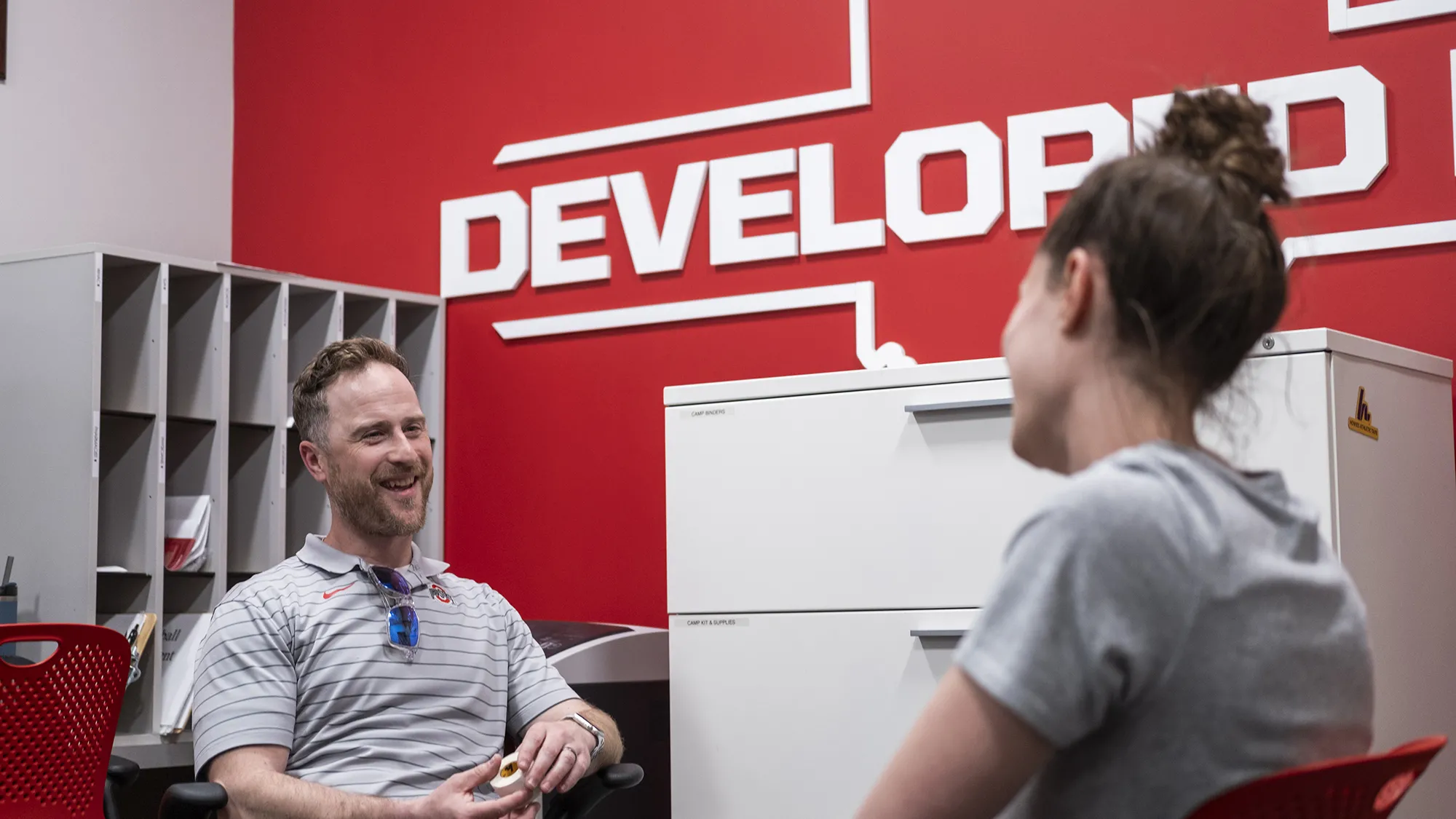 Jamey Houle, a white man in a striped Ohio State golf shirt, laughs as he speaks with a woman shown from the back. They’re both sitting in chairs in an office in the Department of Athletics building. Jamey is a white man with wavy hair and he’s looking directly at his colleague as they chat.