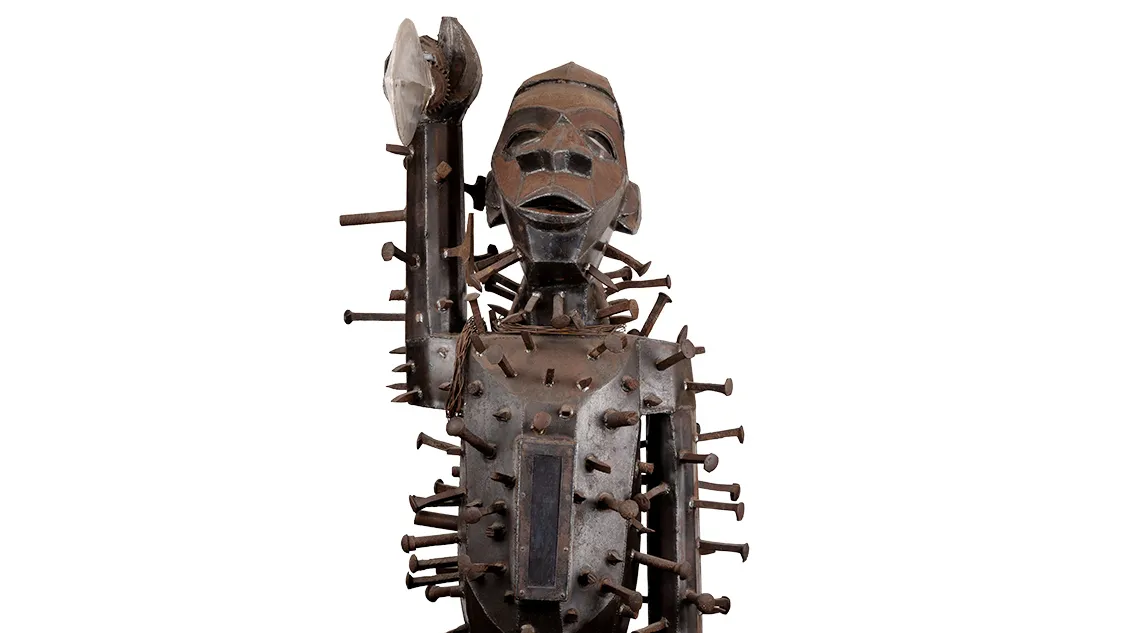 A metal statue with a weathered patina shows an African warrior holding up his sword with thick nails or spikes hammered all over his body. The exception is his face—none is higher than his chin. The warrior looks straight forward. What would usually be rounded—shoulders, belly, arc of the cheek, is rendered in flat panels of metal, adding a geometric look to the warrior. 