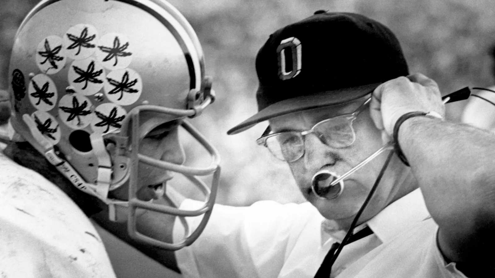 Legendary football coach Woody Hayes stands with Ohio State quarterback Rex Kern, with his arm on his shoulder, as he talks into his headset to players on the field. The image is from Ohio State's archives, and Rex has the trademark buckeye leaf stickers on his helmet, each awarded for an outstanding play.