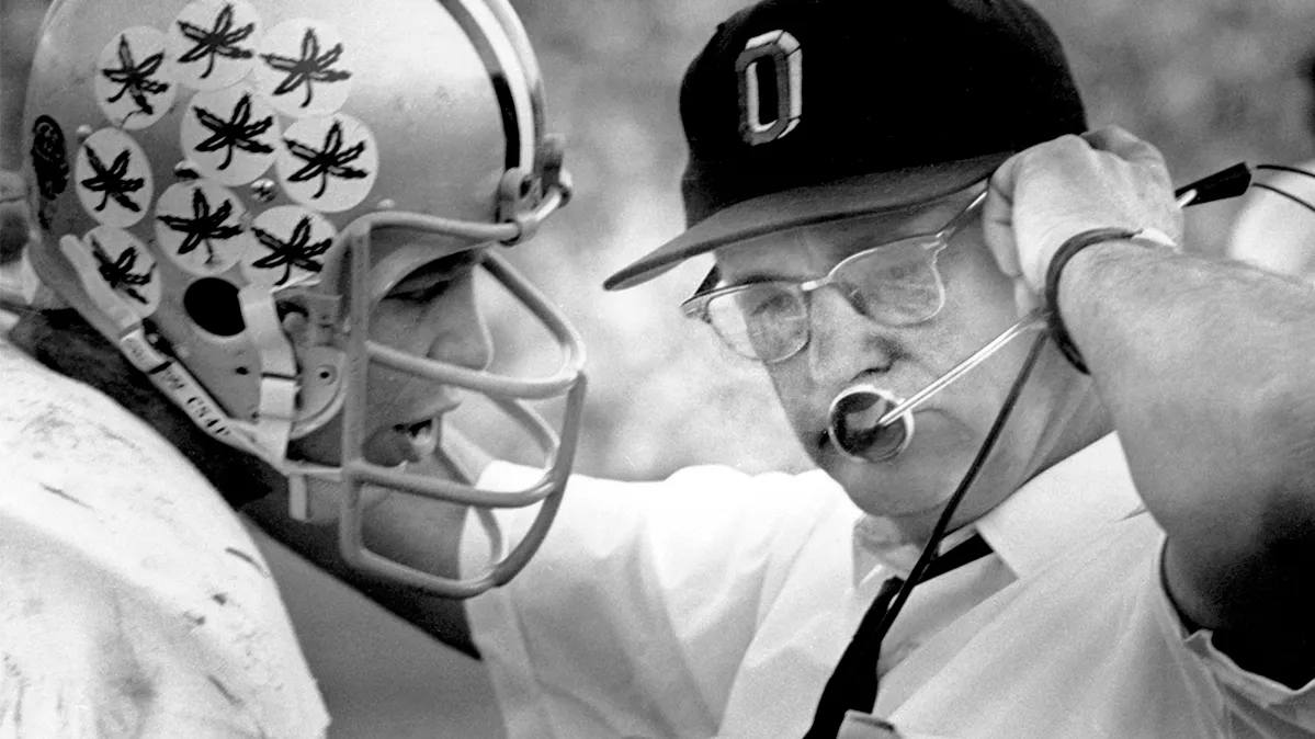 Legendary football coach Woody Hayes stands with Ohio State quarterback Rex Kern, with his arm on his shoulder, as he talks into his headset to players on the field. The image is from Ohio State's archives, and Rex has the trademark buckeye leaf stickers on his helmet, each awarded for an outstanding play.